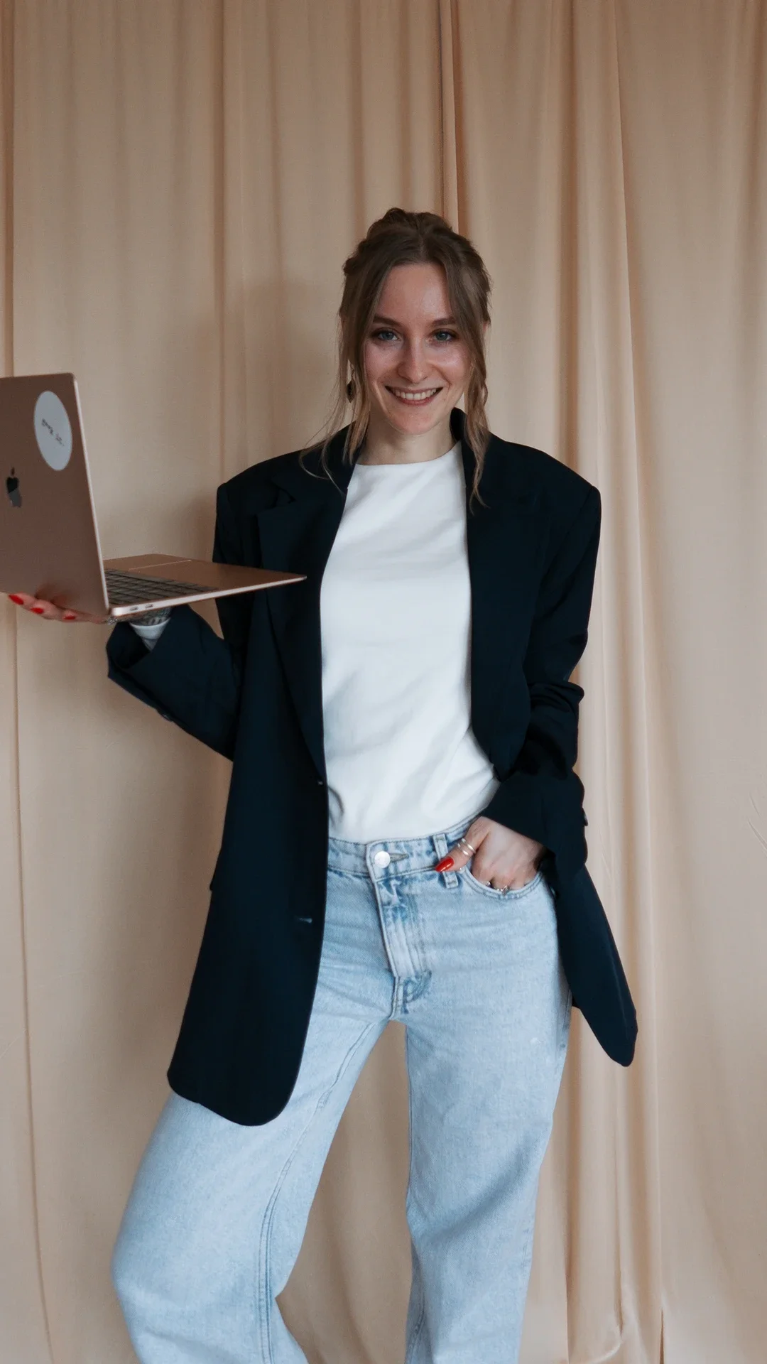 Lea von Macha Branding steht mit einem Laptop in der Hand freundlich lächelnd vor einem hellen Vorhang.