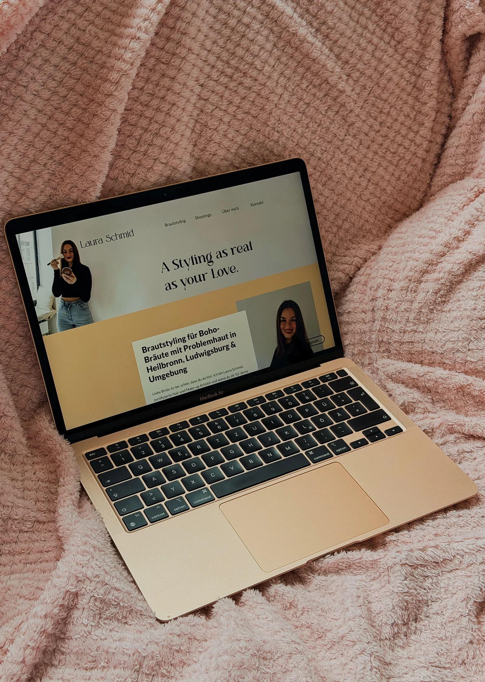 Auf einer rosa Decke liegt ein Laptop, auf dem eine Website geöffnet ist.