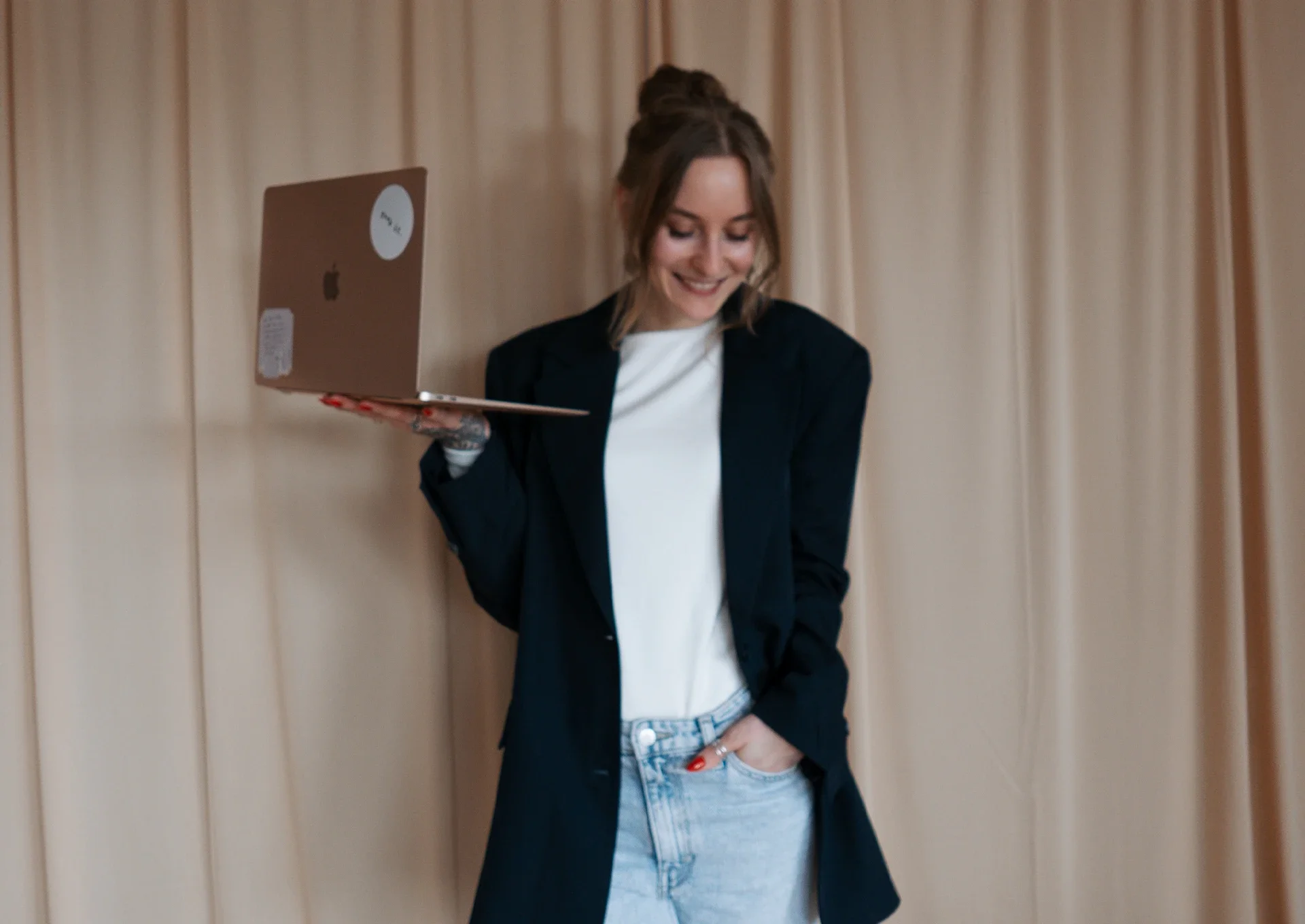Lea steht mit einem Laptop in der Hand vor einem Vorhang und lächelt glücklich.