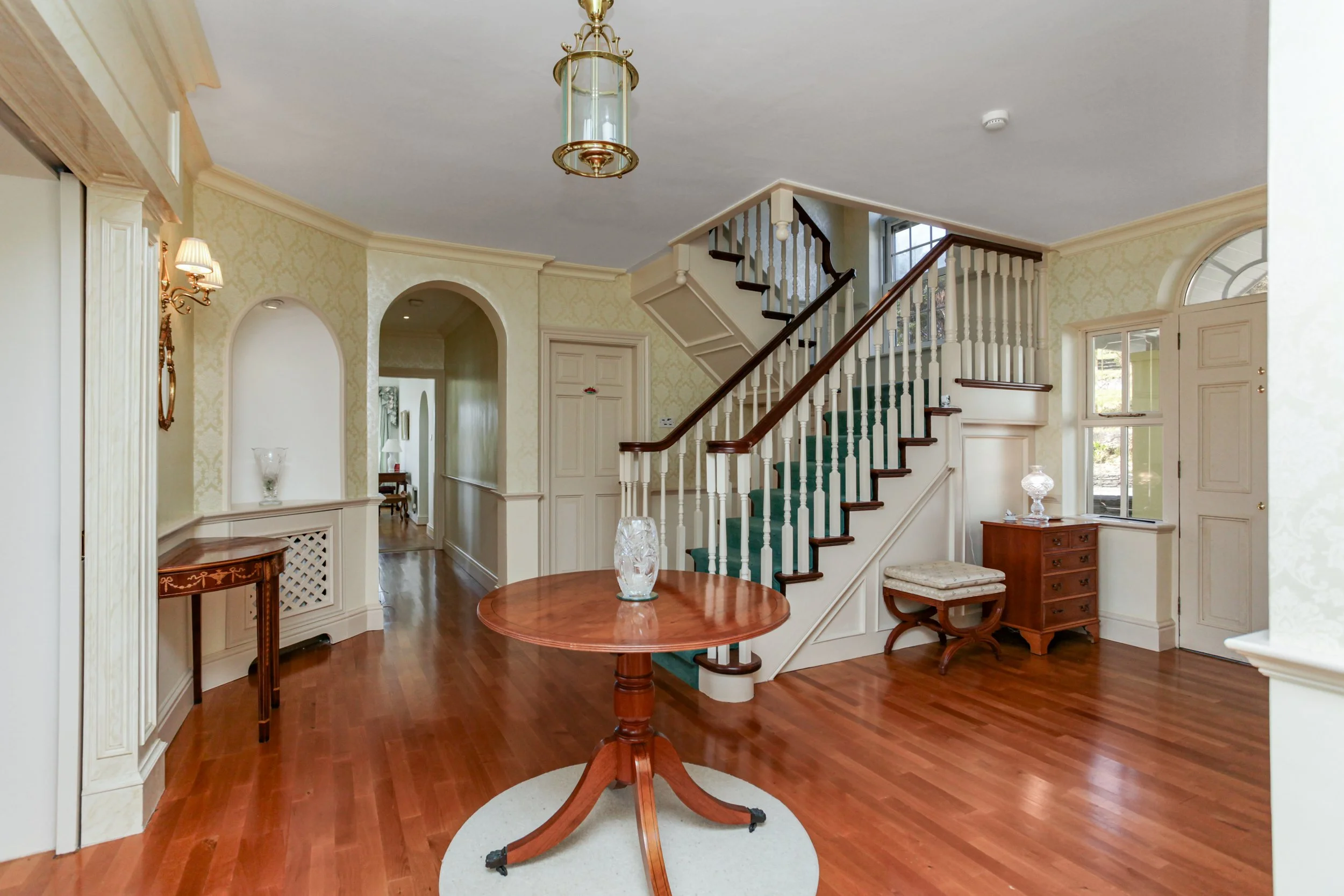 Elegant living room with hardwood floors, a staircase with dark wood handrails and green carpet, and antique wooden furniture, including a round table and a small chest of drawers.