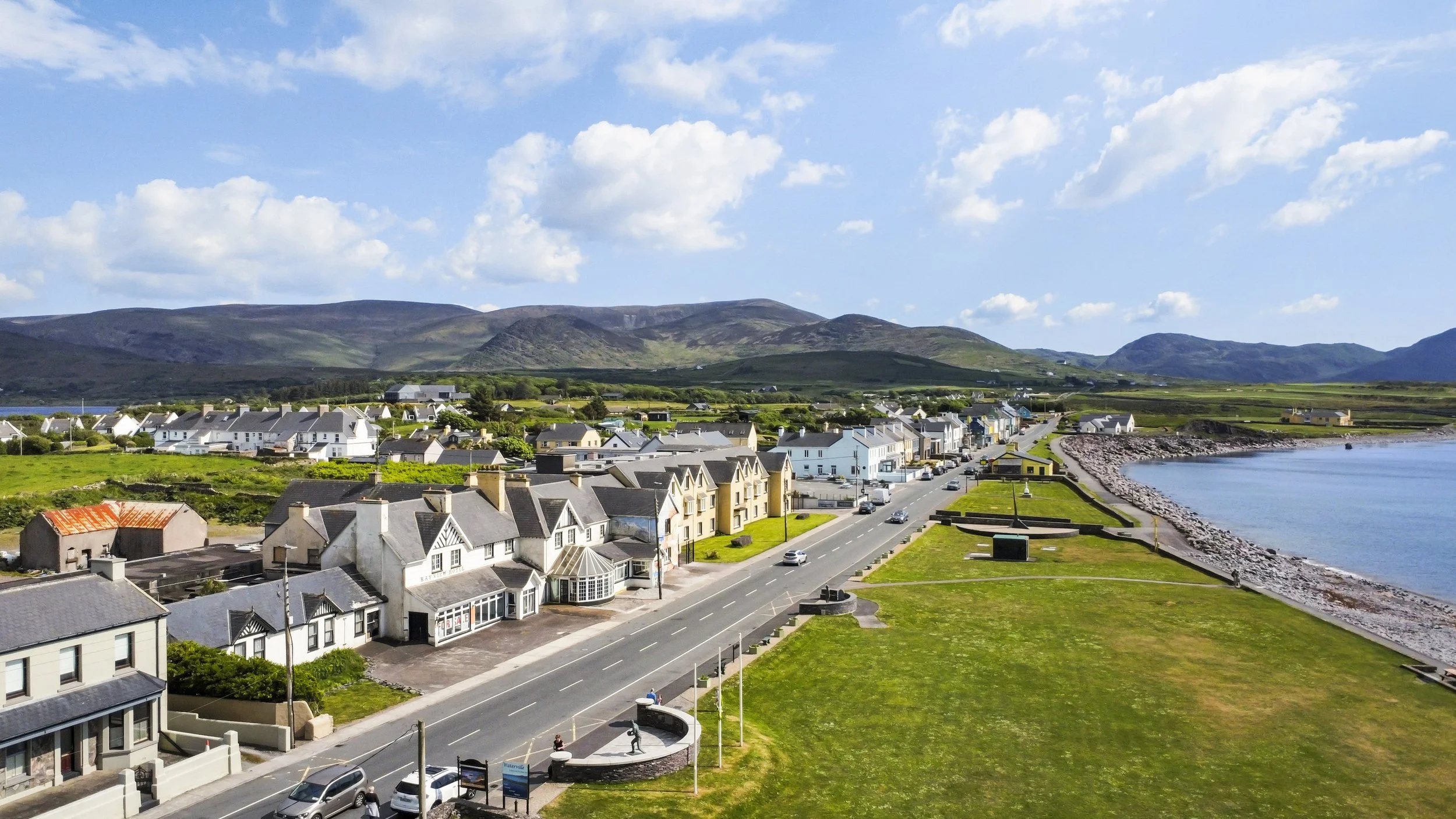 A coastal town with a row of houses along the shoreline, a grassy park, and mountains in the background under a partly cloudy sky.