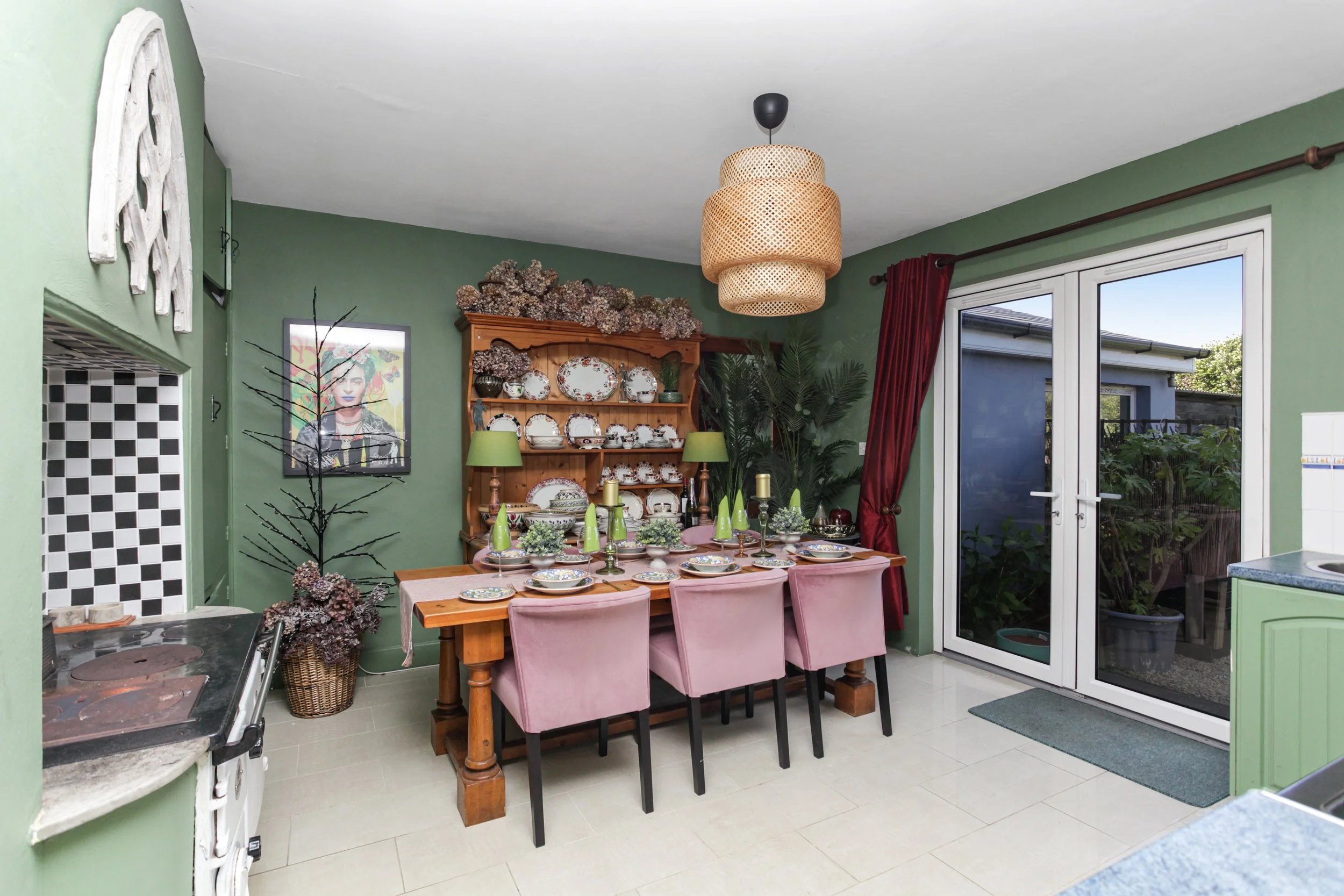 Dining room with green walls decorated with patterned shelves, framed art, and a wooden china cabinet filled with plates and cups. A dining table with pink chairs, set with plates, glasses, and candles. Large sliding glass door with red curtains lead