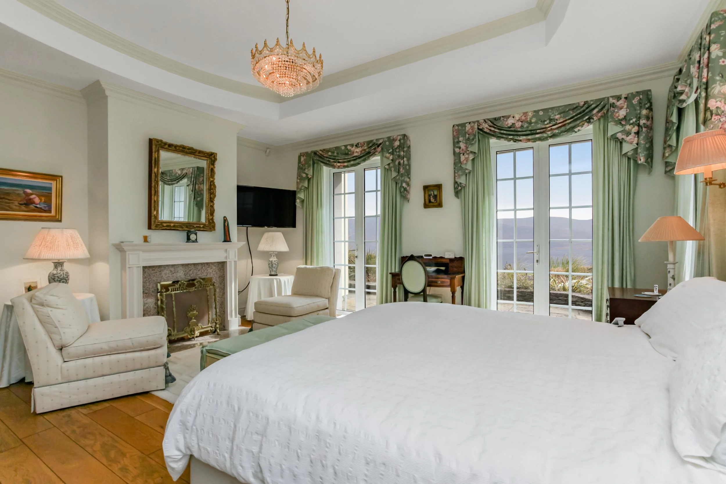 A spacious bedroom with a large bed, green floral curtains, a fireplace, a television, a small writing desk, and scenic outdoor view