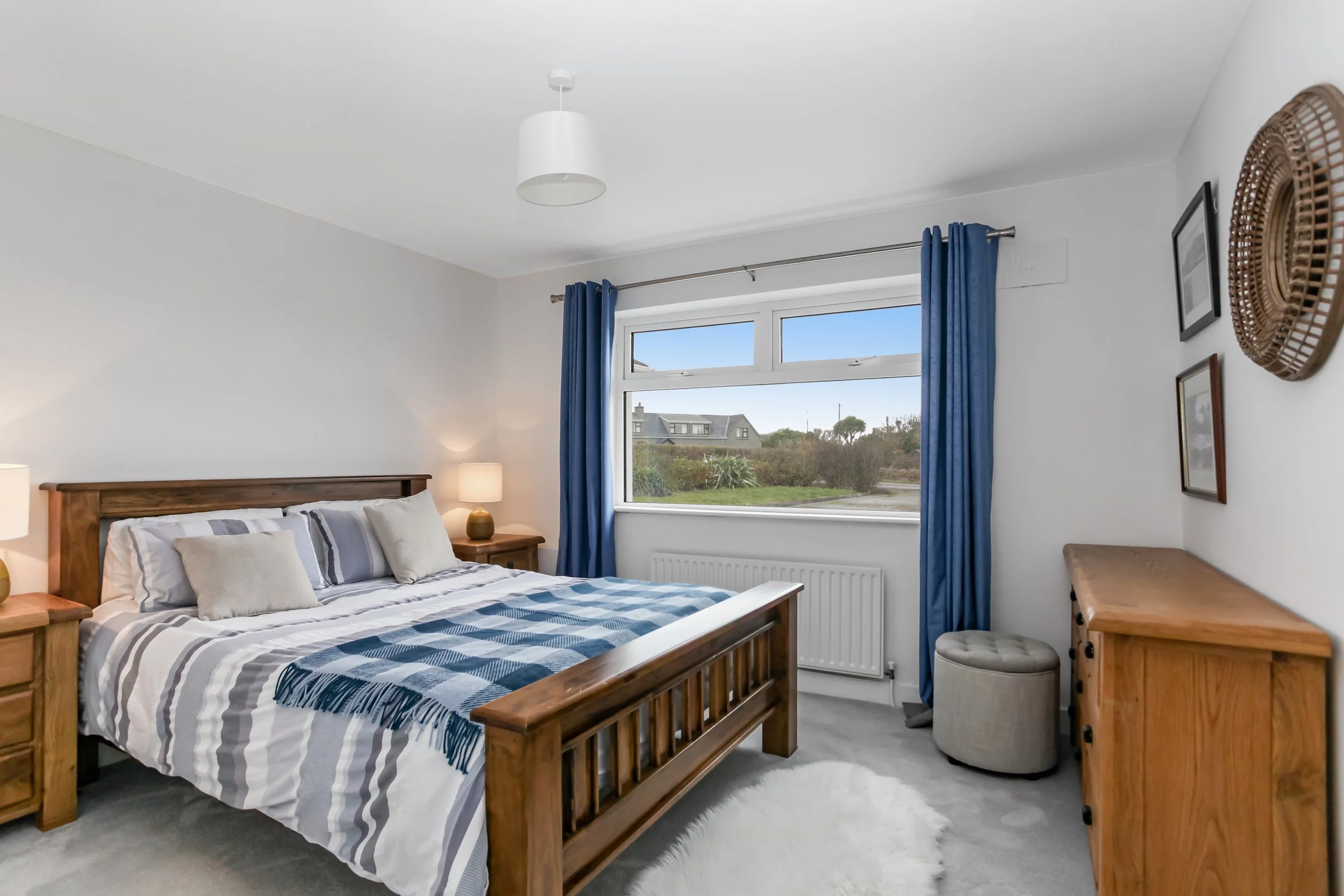 A bedroom with a wooden bed frame, matching bedside tables with lamps, a large window with blue curtains showing an outside view of houses and greenery, a wooden dresser, framed pictures on the wall, a beige ottoman, and a fluffy white rug.