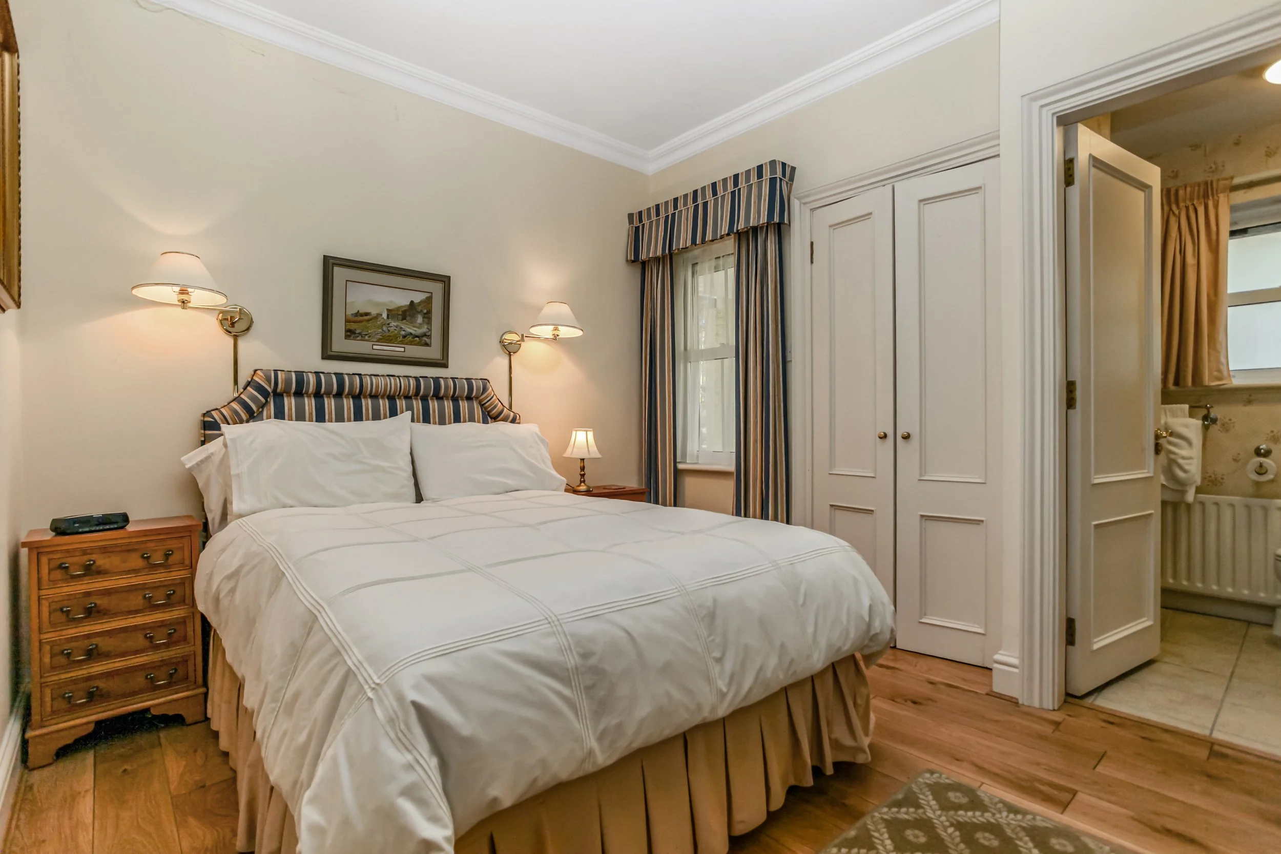 A bedroom with a bed, wooden nightstands, and a closet. The bed has white bedding and a striped headboard. There are windows with striped curtains. A bathroom is partially visible through an open door.