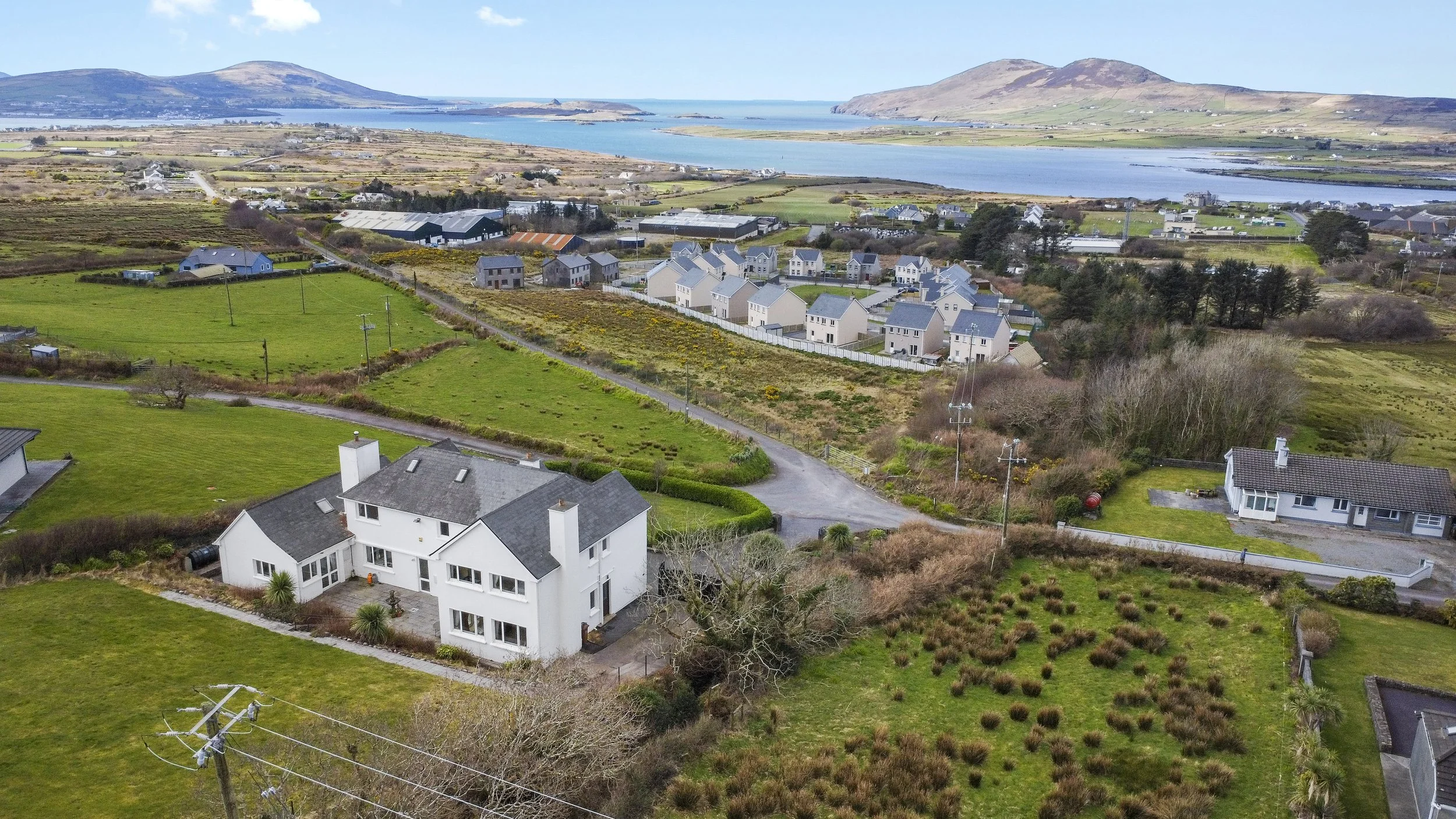 Aerial view of a coastal residential neighborhood with houses, green lawns, and fields, overlooking a body of water with hills in the background.