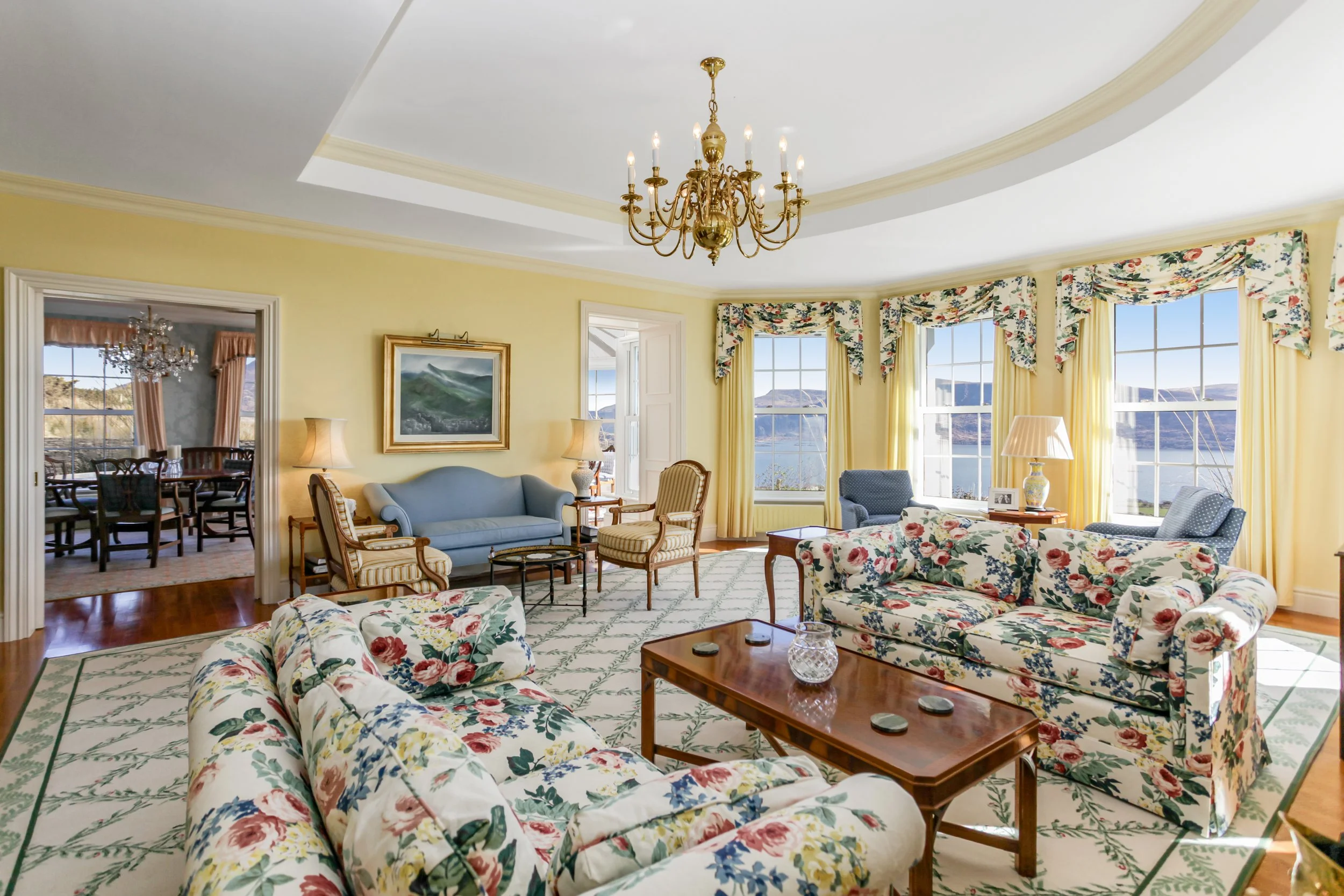 Bright living room with large bay windows, floral and pastel furniture, a chandelier, and a scenic mountain view outside.