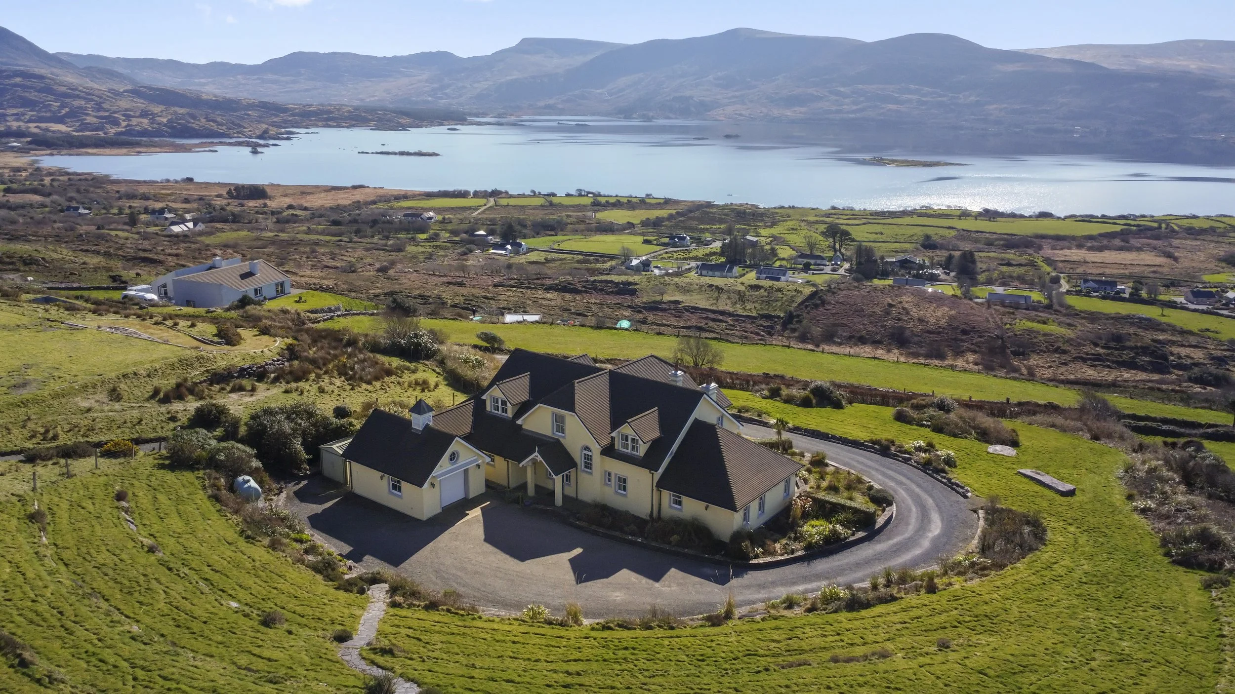 A large yellow house with multiple gabled roofs and a circular driveway, situated on a lush green hillside overlooking a lake and mountains in the distance.