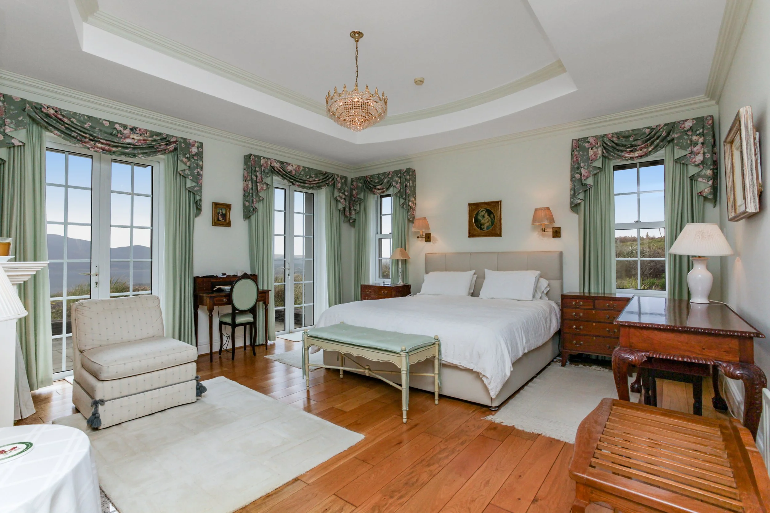 A spacious bedroom with a large bed, multiple windows with floral valances and green curtains, hardwood floors, and vintage-style furniture including dressers, a sitting bench at the foot of the bed, a small desk with a chair, and a cushioned armchai
