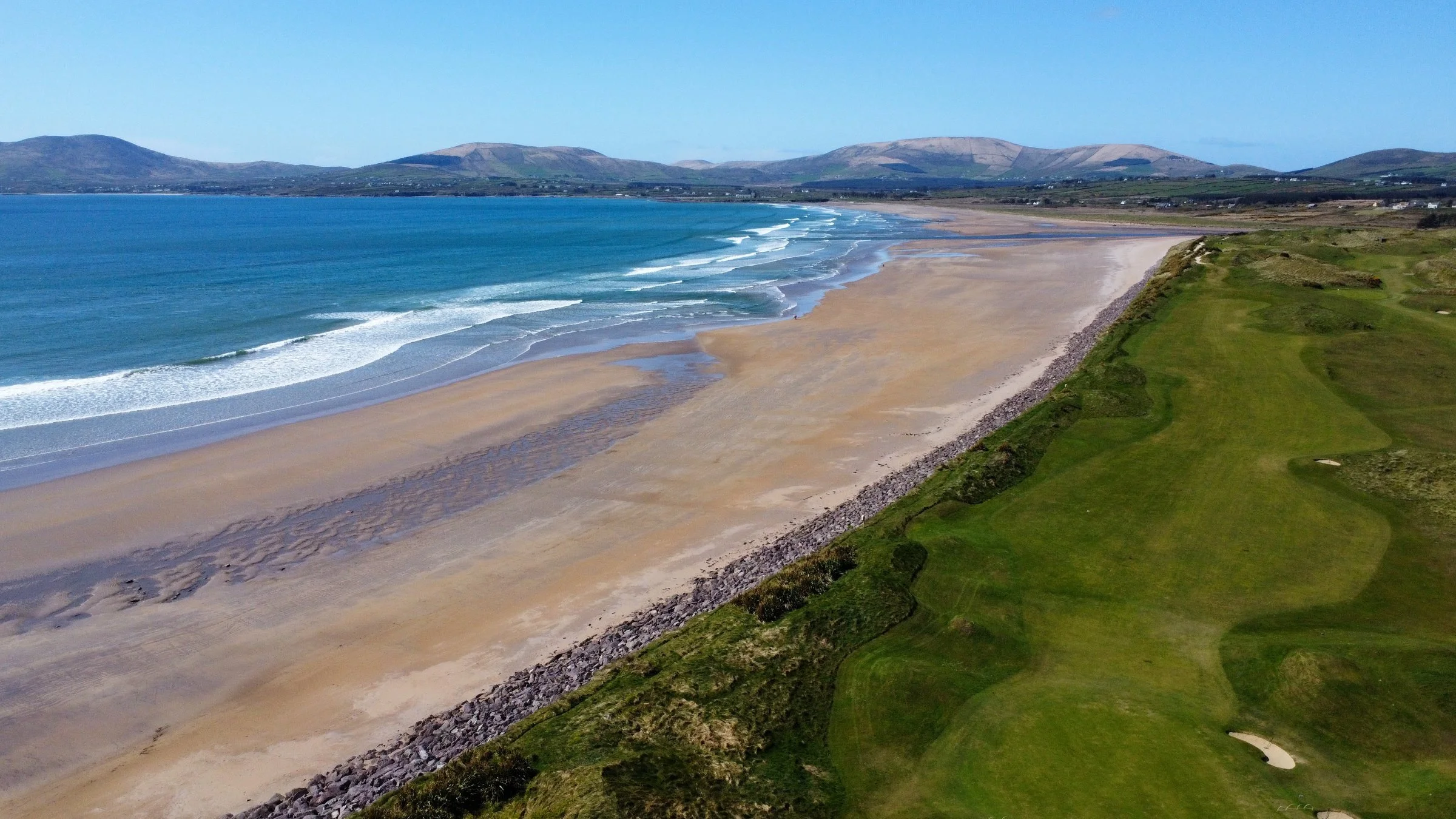 Aerial view of a sandy beach with waves, green grass area, and hills in the background under a clear blue sky.