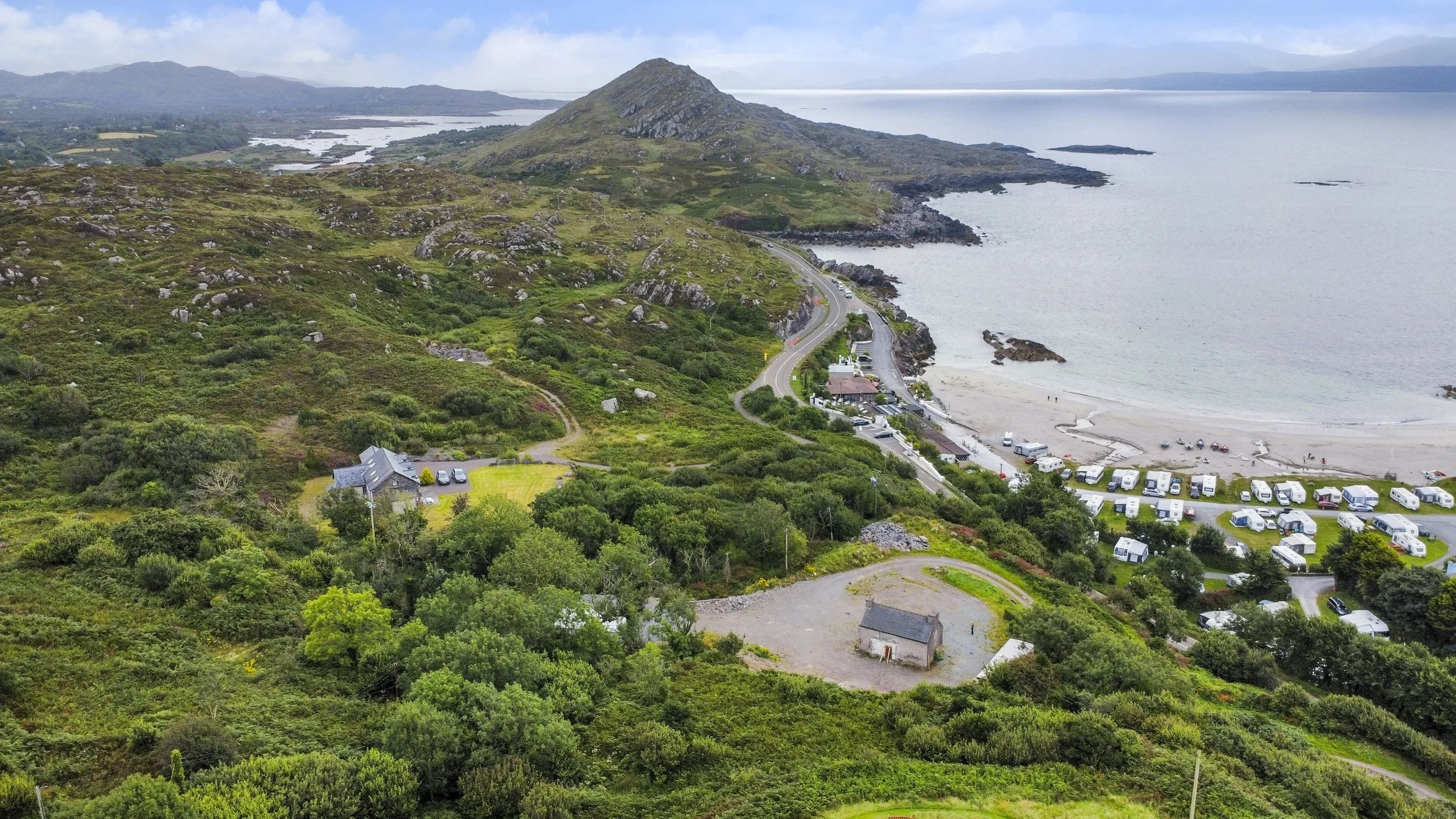 Aerial view of a coastal landscape with a road, a beach, and green hills with scattered buildings and caravans near the shoreline.