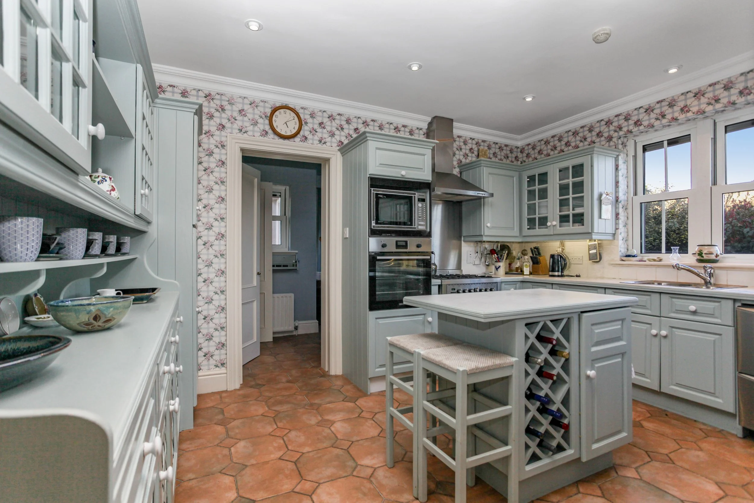 A traditional kitchen with light blue cabinets, terracotta tile flooring, and floral wallpaper. Features include a kitchen island with wine storage, built-in oven and microwave, and a window above the sink.