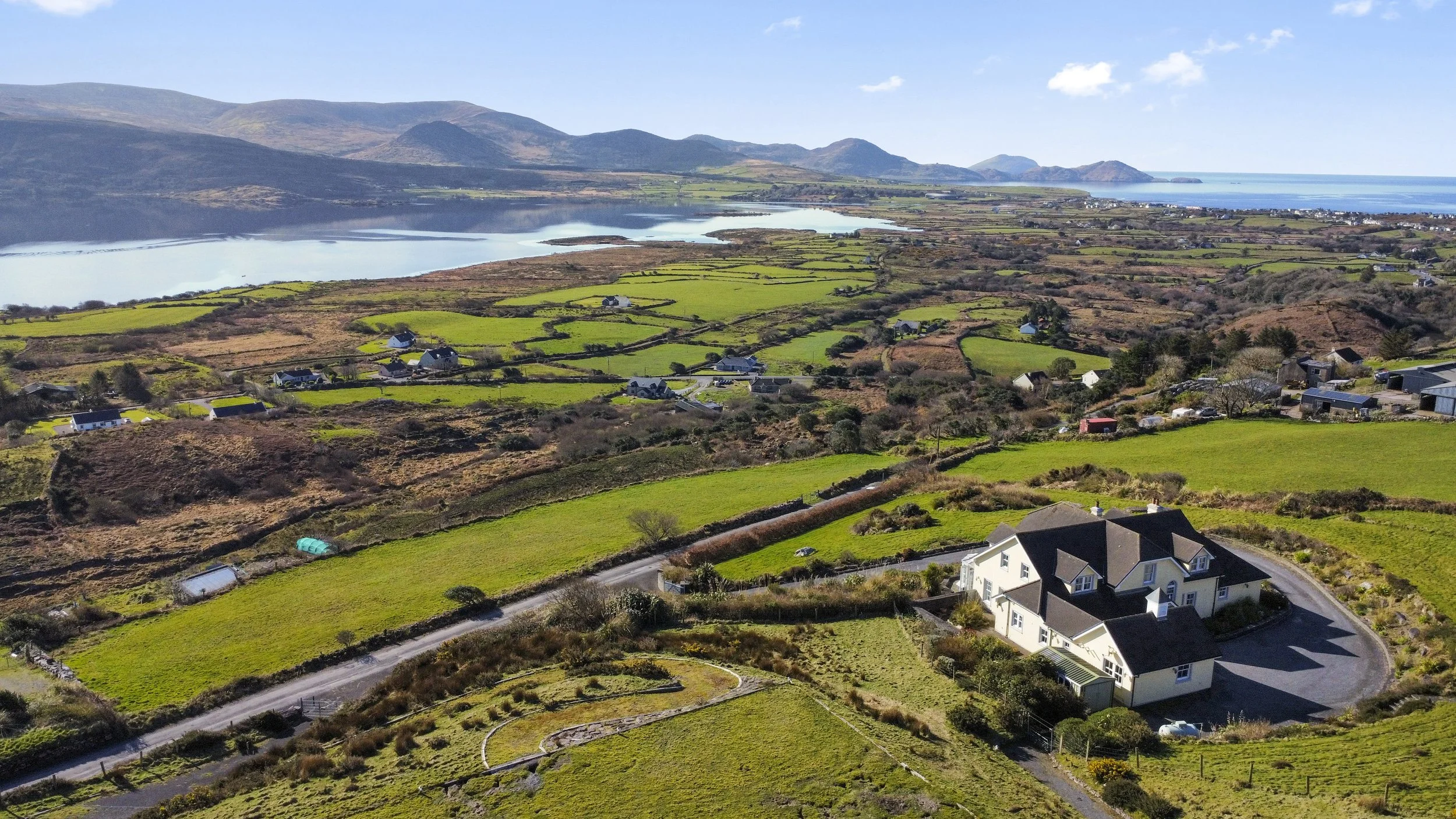 Aerial view of a rural landscape with a large house, green fields, scattered houses, a lake, and distant mountains on a sunny day.
