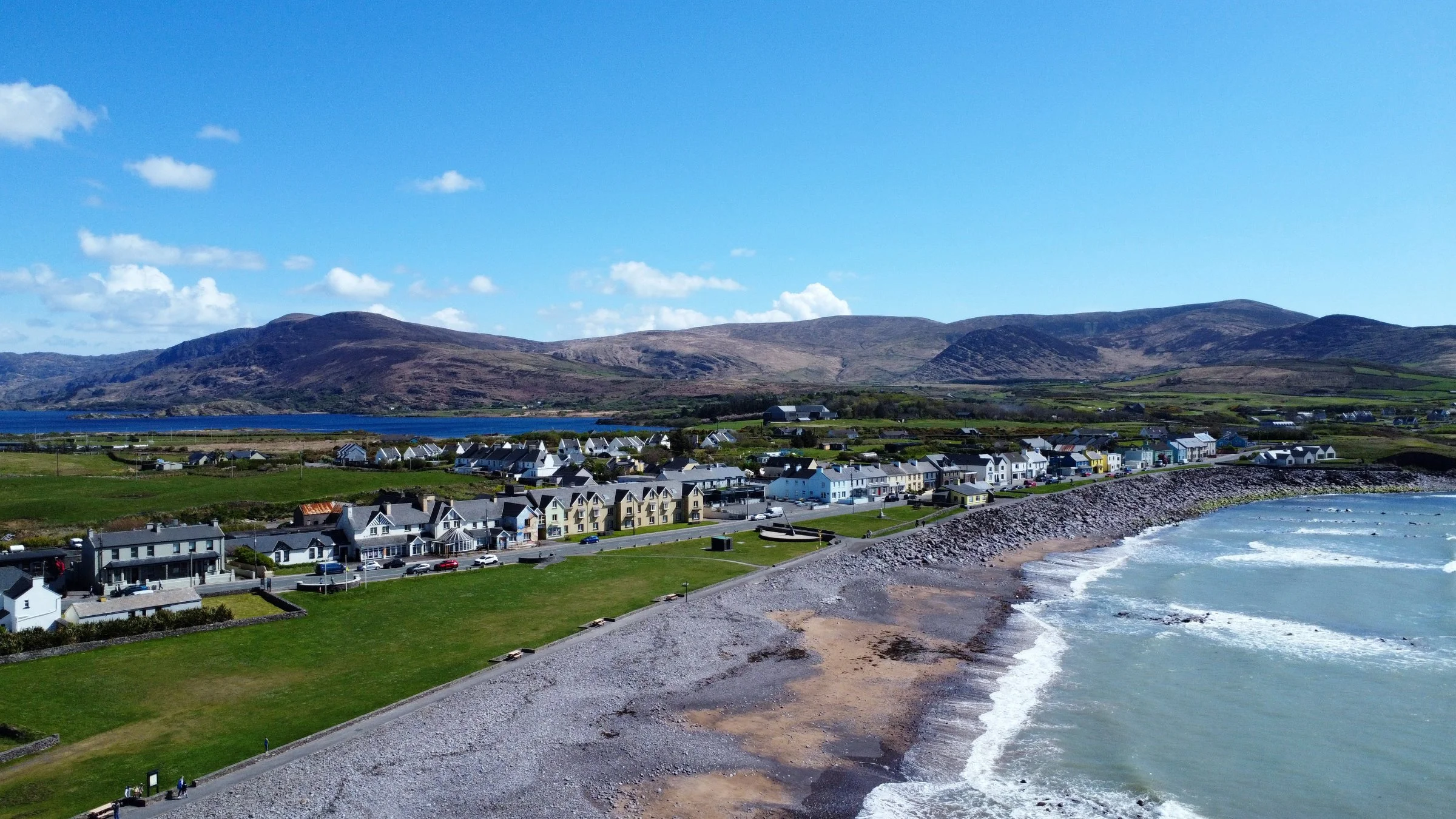 A coastal village with colorful houses along a pebble beach, green fields, hills, and a blue sky with scattered clouds.