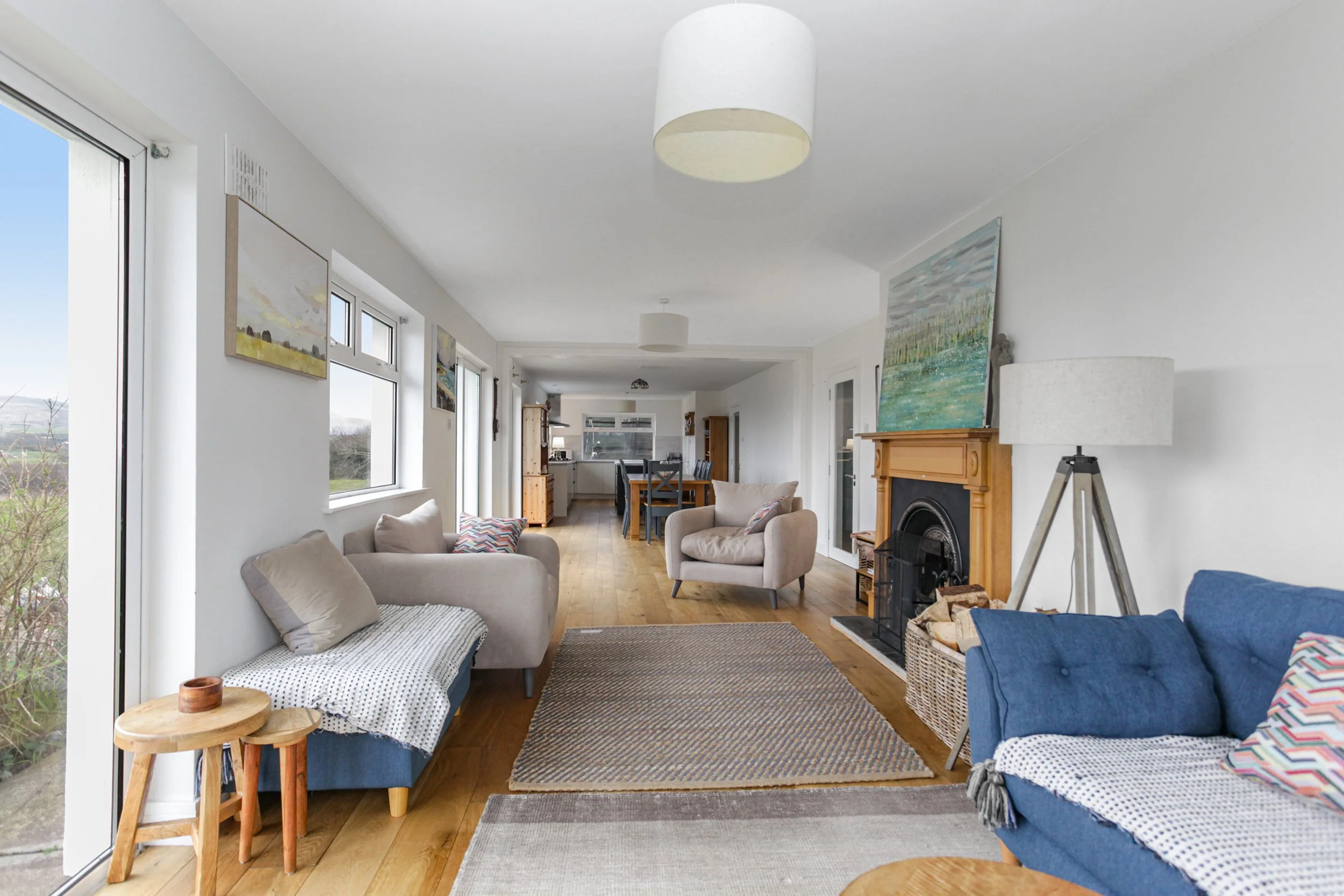 Living room with beige and blue sofas, a fireplace with a wooden mantel, paintings on the wall, a tripod floor lamp, a window seat, and dining area in the background.