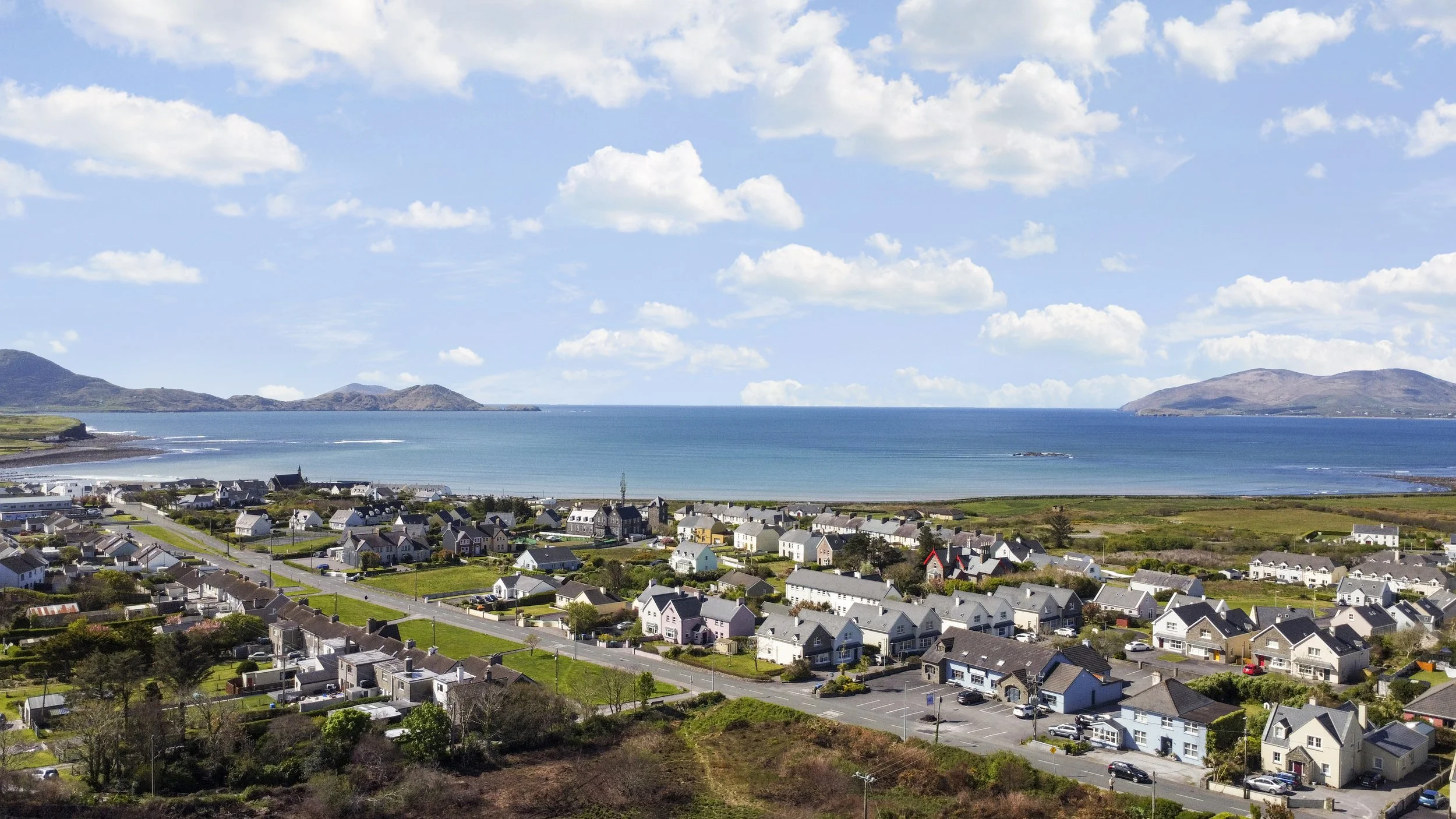 A coastal town with colorful houses, a beach, and a calm sea, surrounded by hilly landscape and a partly cloudy sky.