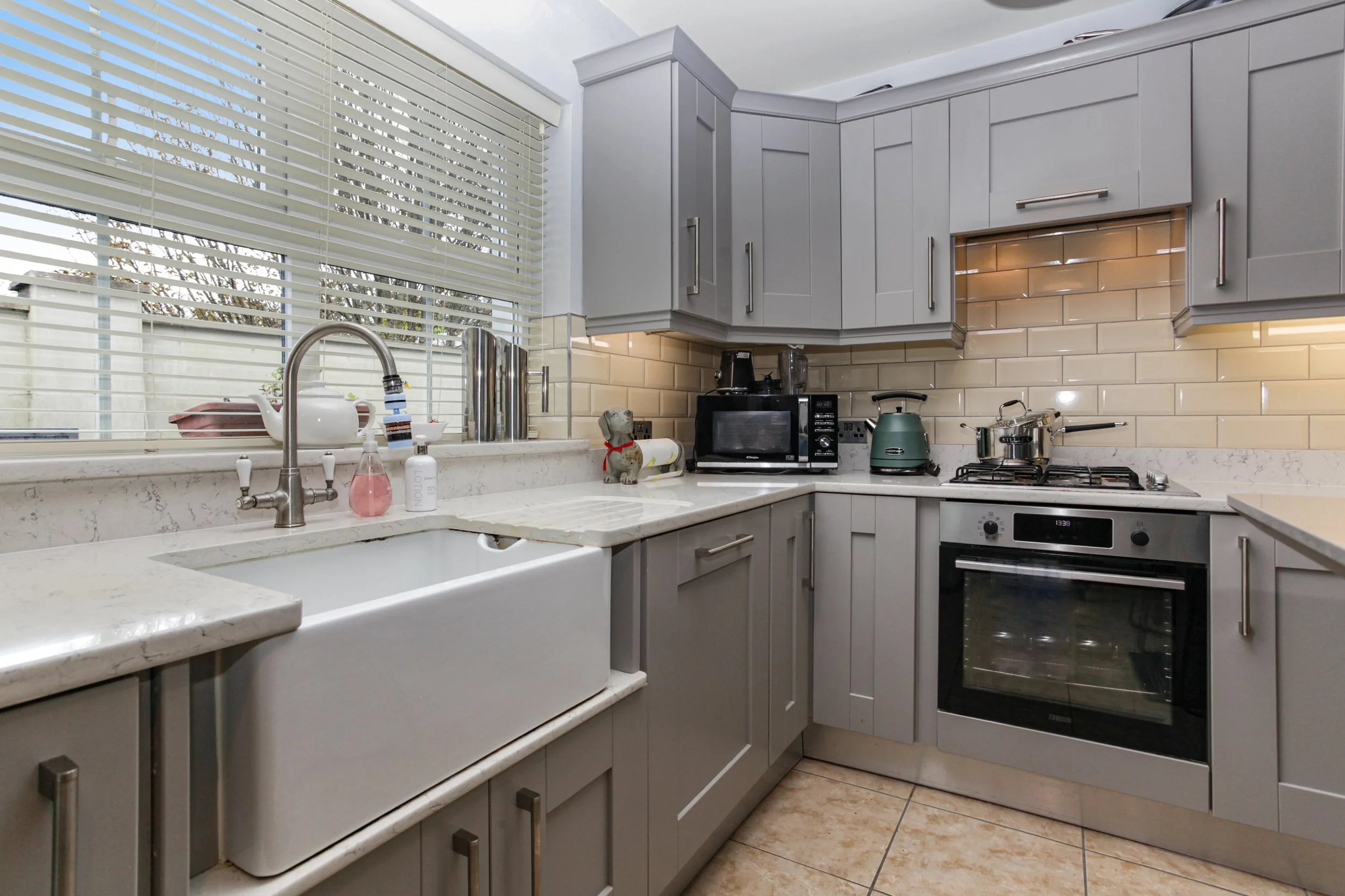 Kitchen with gray cabinets, beige backsplash, and a white farmhouse sink under a window with blinds, with appliances like a microwave, kettle, and stove.