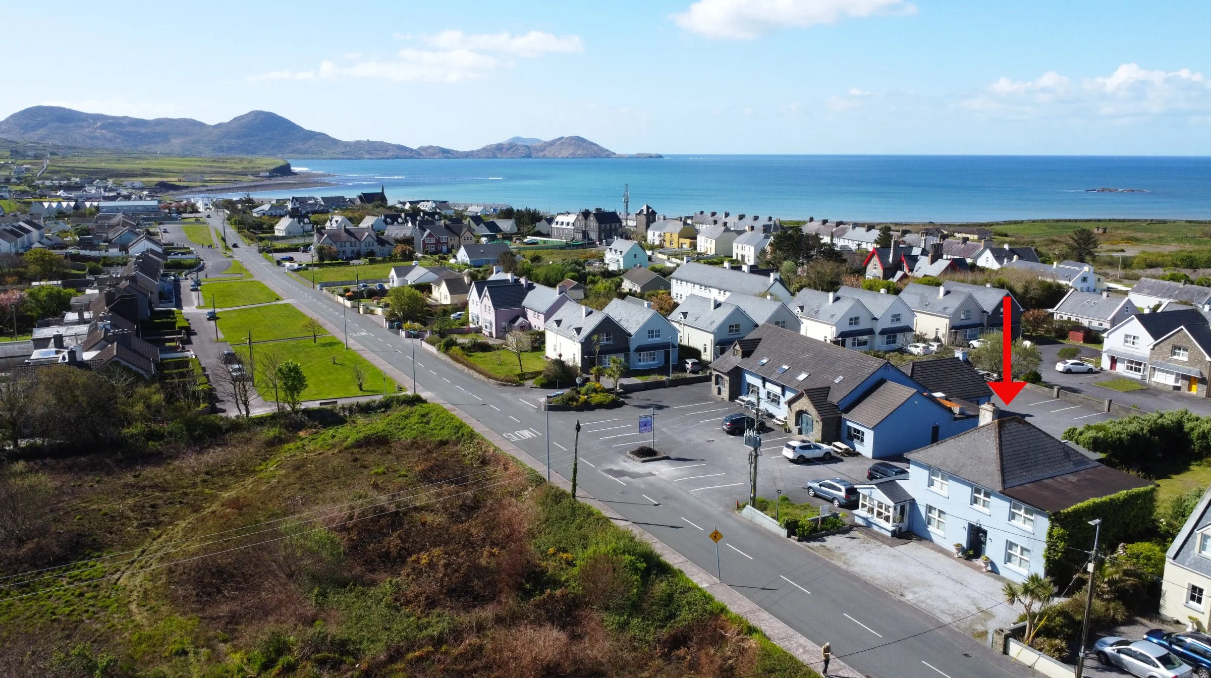 Aerial view of a coastal town with houses, roads, and parking lots near the ocean, with hills and the sea in the background.