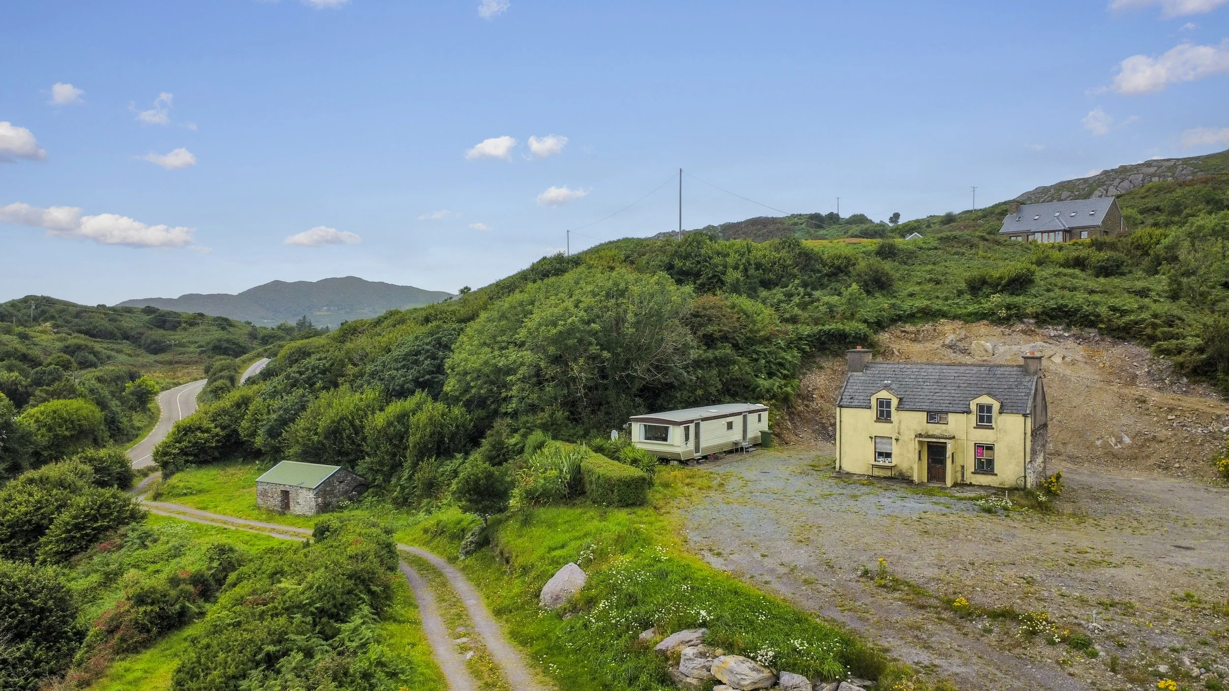 A rural landscape with a winding road, a small stone building, a caravan, and a yellow house on a hillside surrounded by greenery and rolling hills under a blue sky with scattered clouds.