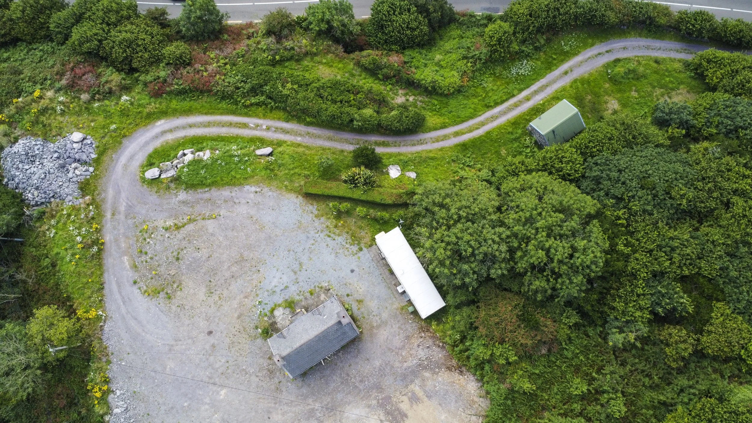 An aerial view of a rural property with a gravel driveway, a small house, a large shed, and surrounding lush green trees and bushes.