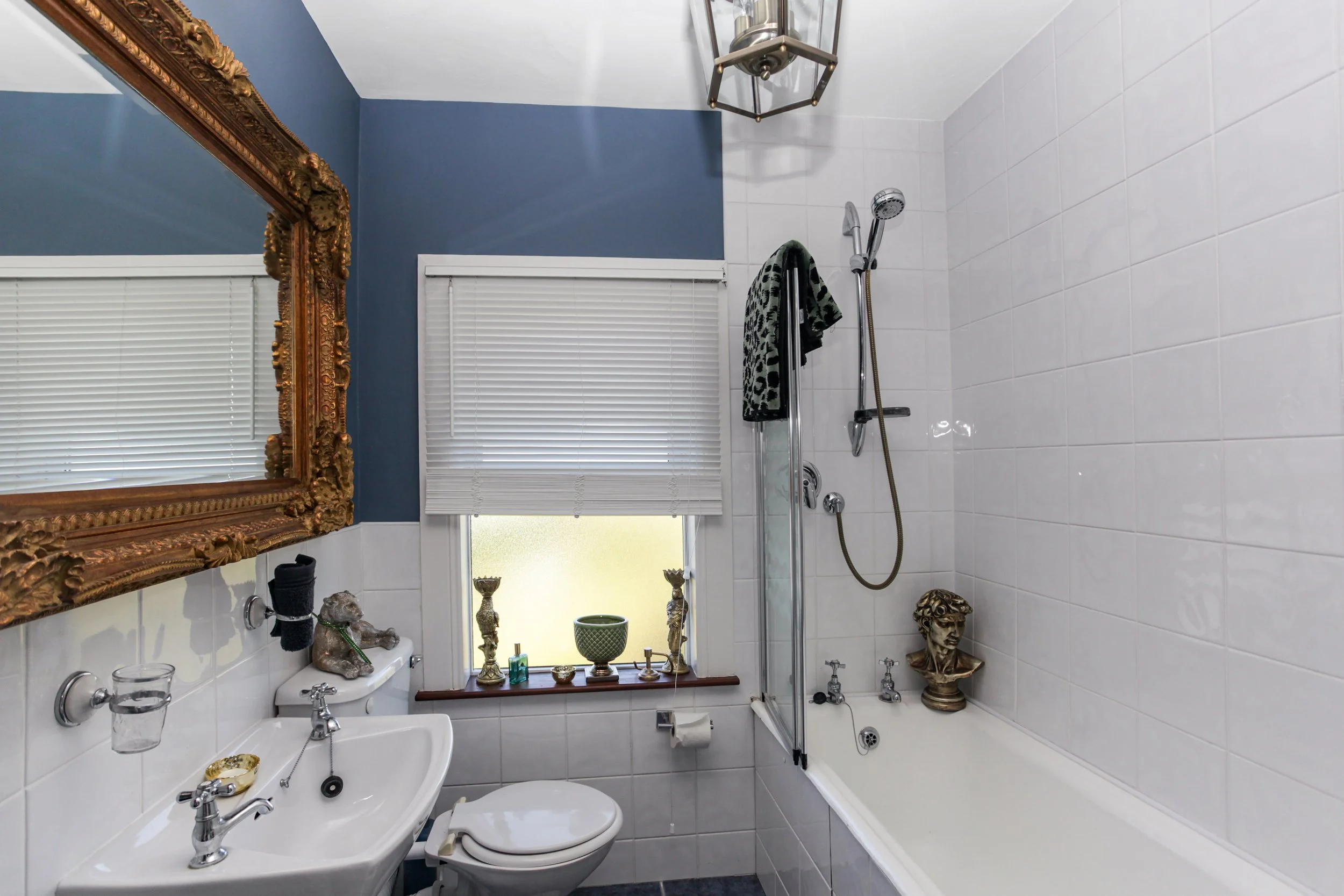 Decorative bathroom with gold-framed mirror, blue and white walls, small sink, toilet, window with blinds, bathtub with showerhead, and various decorative sculptures and objects on windowsill and bathtub edge.