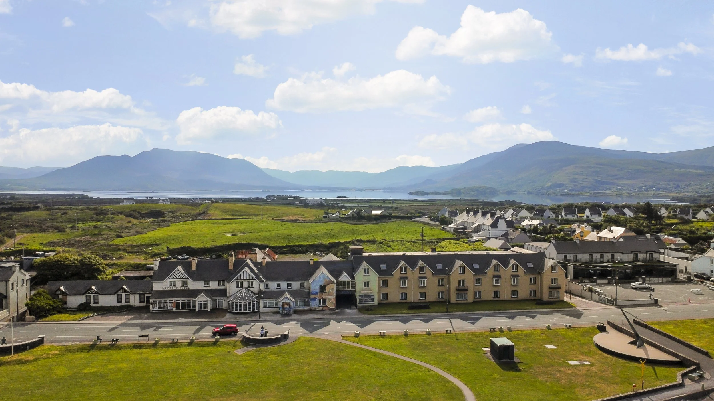 View of a town with colorful buildings, green fields, a large grassy park, and mountains in the distance under a partly cloudy sky.