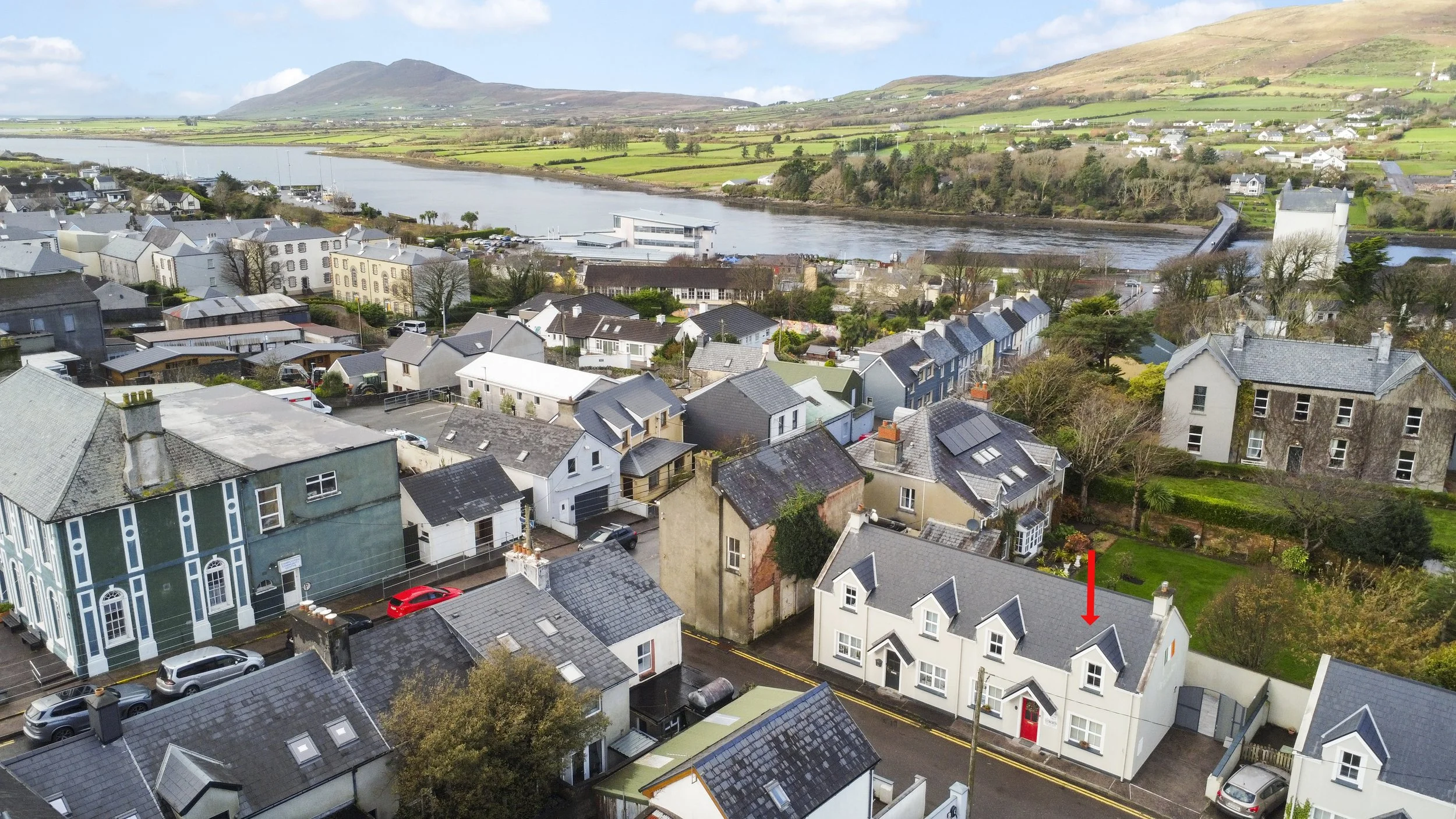 Aerial view of a small town with houses, cars on the street, a river on the edge of town, bridge crossing the river, and green hills in the background.