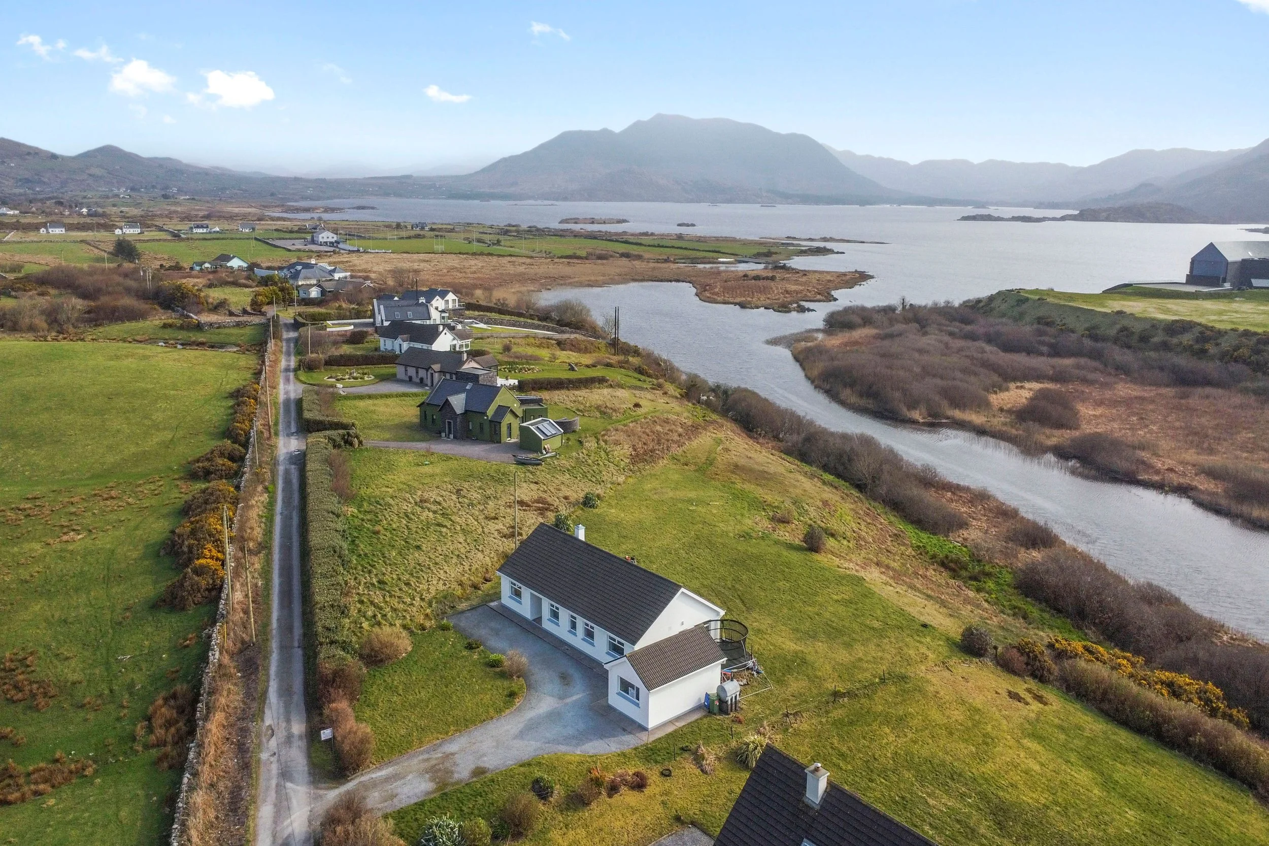 Aerial view of a rural neighborhood near a body of water, with houses, green fields, and a winding river, surrounded by mountains in the background.