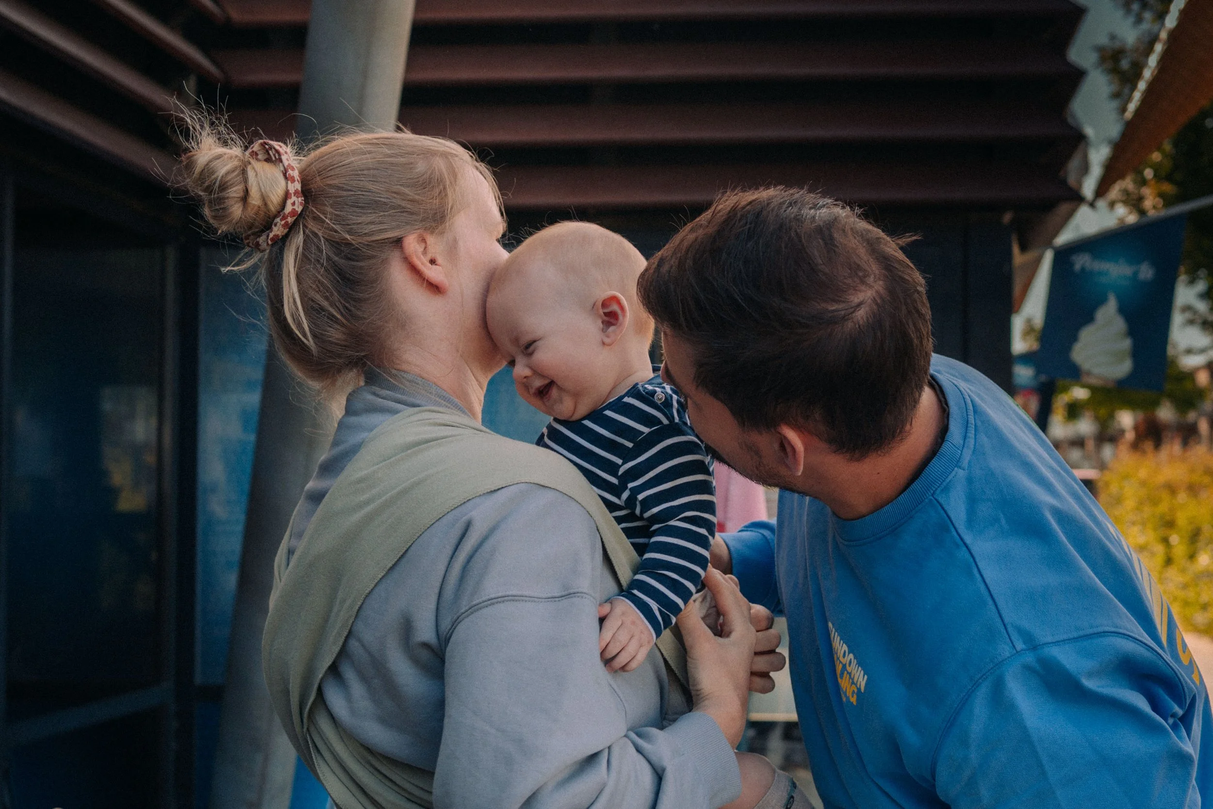 Ein Baby wird von einer Mutter und einem Vater auf einem Jahrmarkt begrüßt, alle lachen und haben Spaß.