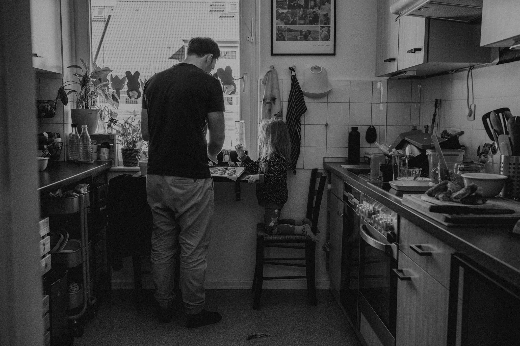 Ein Mann und ein kleines Mädchen stehen in einer Küche, das Mädchen sitzt auf einem Stuhl, der Mann steht vor ihr, beide schauen auf eine Arbeitsfläche. Die Küche ist voll mit Küchengeräten und -utensilien, es gibt Pflanzen auf der Fensterbank und an