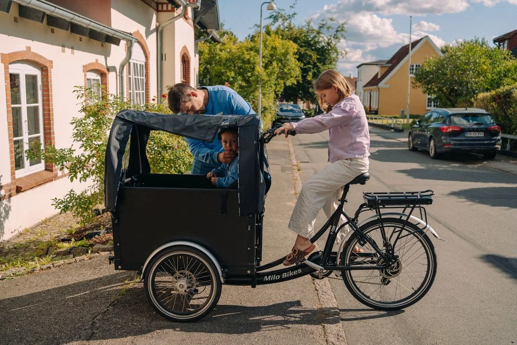 Ein Mädchen fährt mit einem Fahrrad mit Anhänger, in dem ein Kind sitzt, während ein erwachsener Mann im Hintergrund steht.