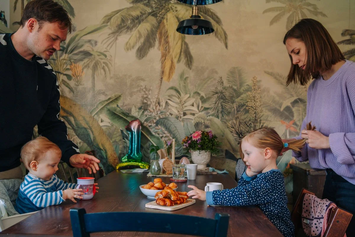 Familie mit zwei Kindern bei einem Frühstückstisch, auf dem Croissants, Kaffee und Heißgetränke stehen, in einer gemütlichen, pflanzenreichen Küche