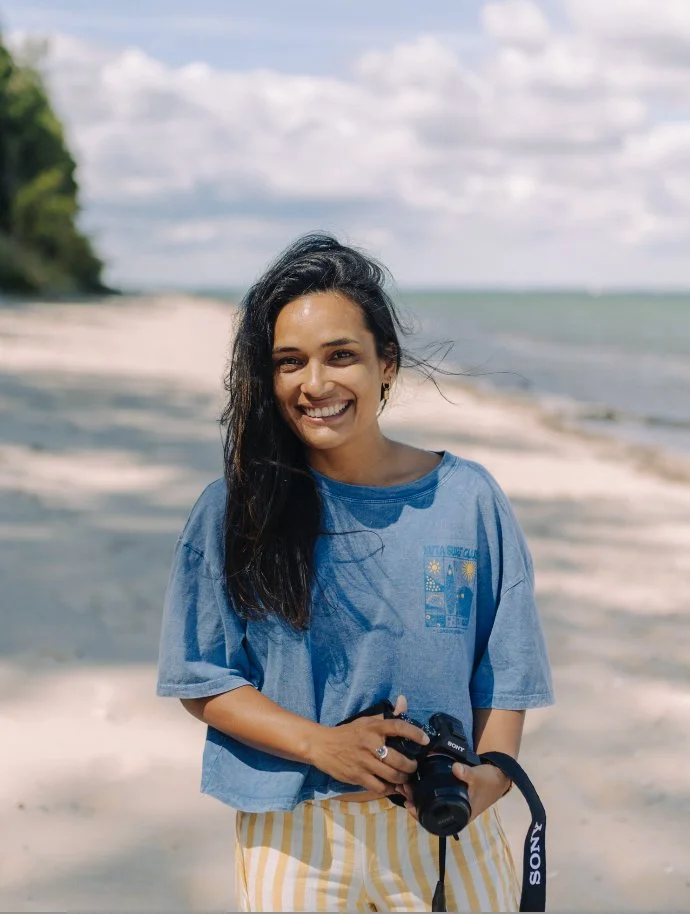 Lächelnde Frau mit Kamera am Strand, im Hintergrund Meer und Himmel mit Wolken.
