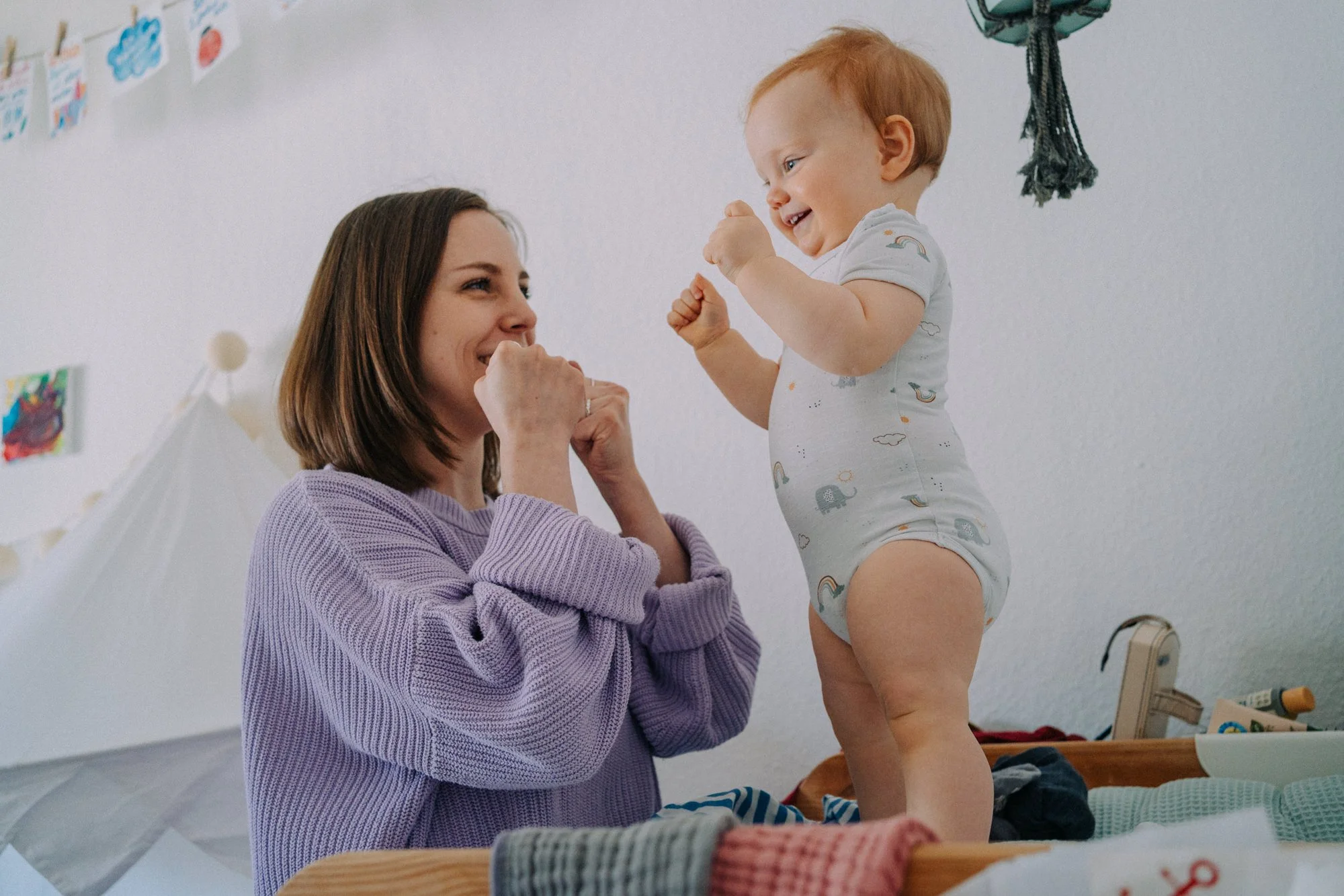 Eine Frau hebt eine fröhliche, lachende Baby an, während sie miteinander spielen. Die Szene ist in einem hellen Raum mit Kinderbildern an der Wand.
