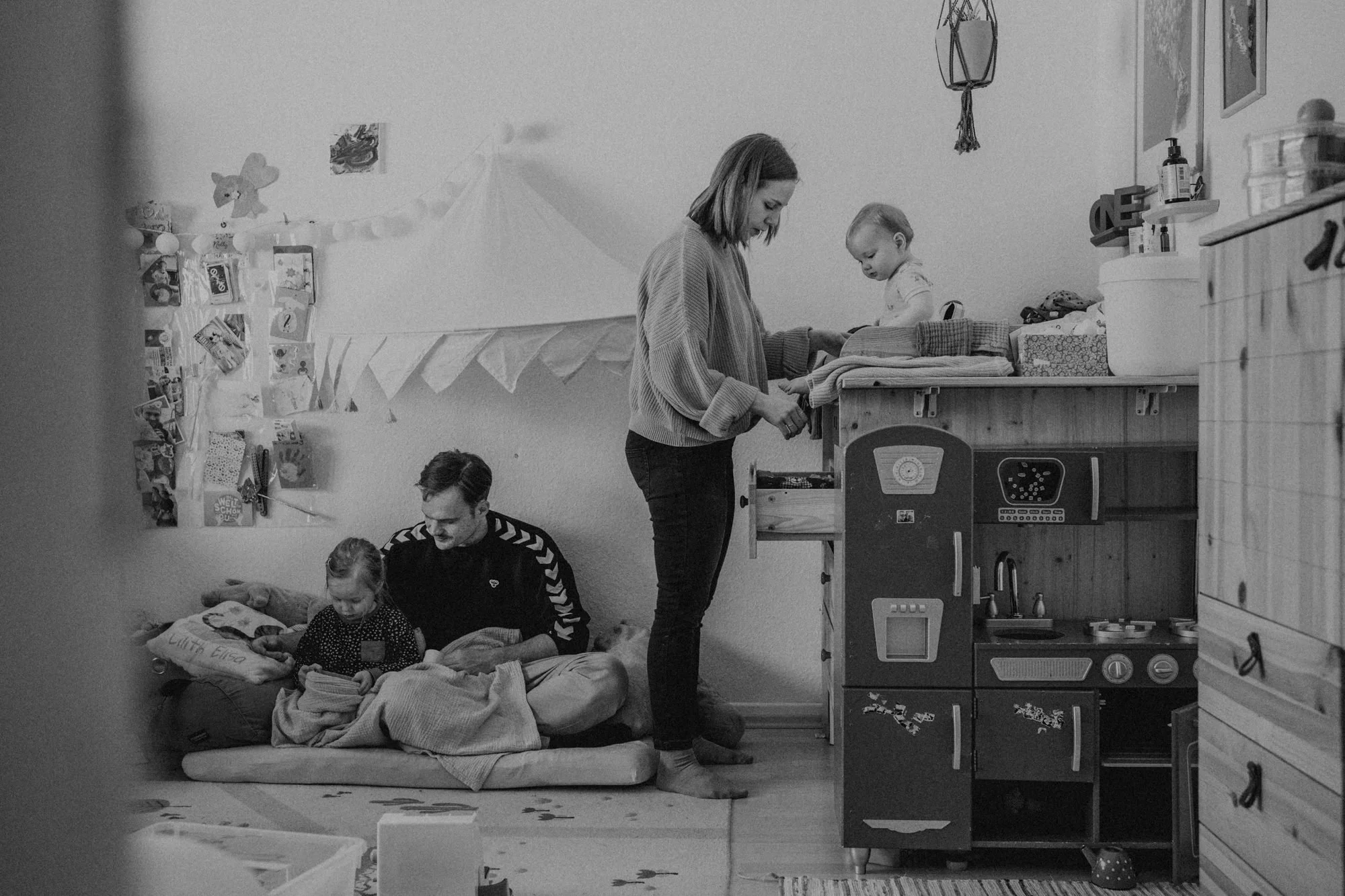 Familie mit Kindern im Wohnzimmer, ein Kind sitzt auf einem Erwachsenen im Bett und schaut auf ein Tablet, während eine Frau beim Wickeln eines Kindes auf einer Wickelkommode ist.