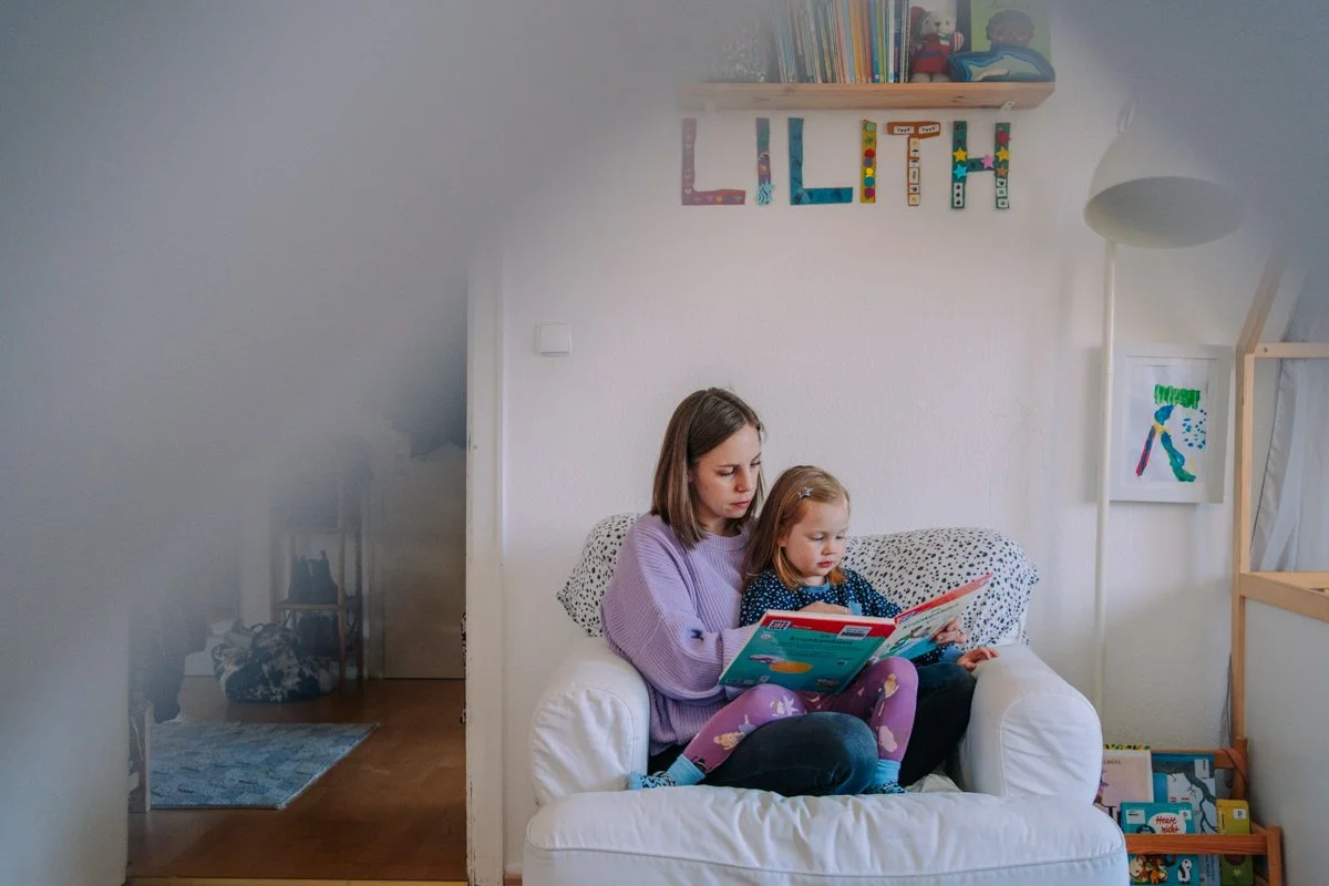 Eine Frau liest mit einem Mädchen auf einem weißen Sessel in einem Kinderzimmer.