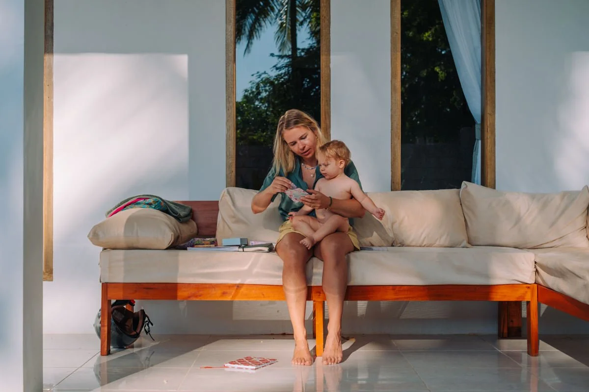 Eine Frau sitzt auf einem Sofa und hält ein Baby auf dem Schoß. Sie schauen gemeinsam in eine Zeitschrift. Das Wohnzimmer ist hell erleuchtet mit offenen Fenstern und Blick ins Freie.