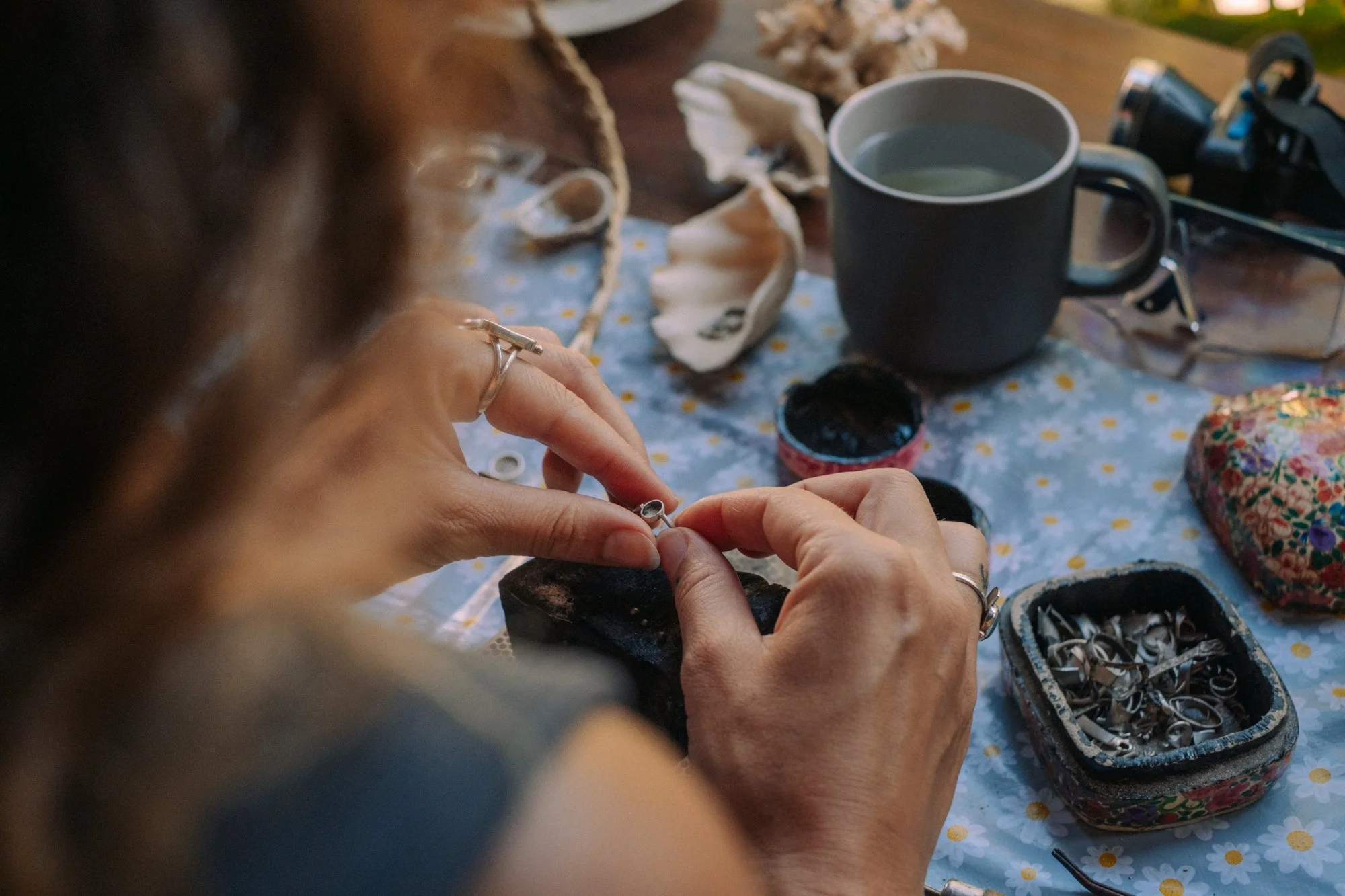 Person arbeitet an Schmuckstücken mit kleinen Metallringen, umgeben von Werkzeugen, Schmuckmaterialien und einer schwarzen Tasse mit Getränk auf einem Tisch.
