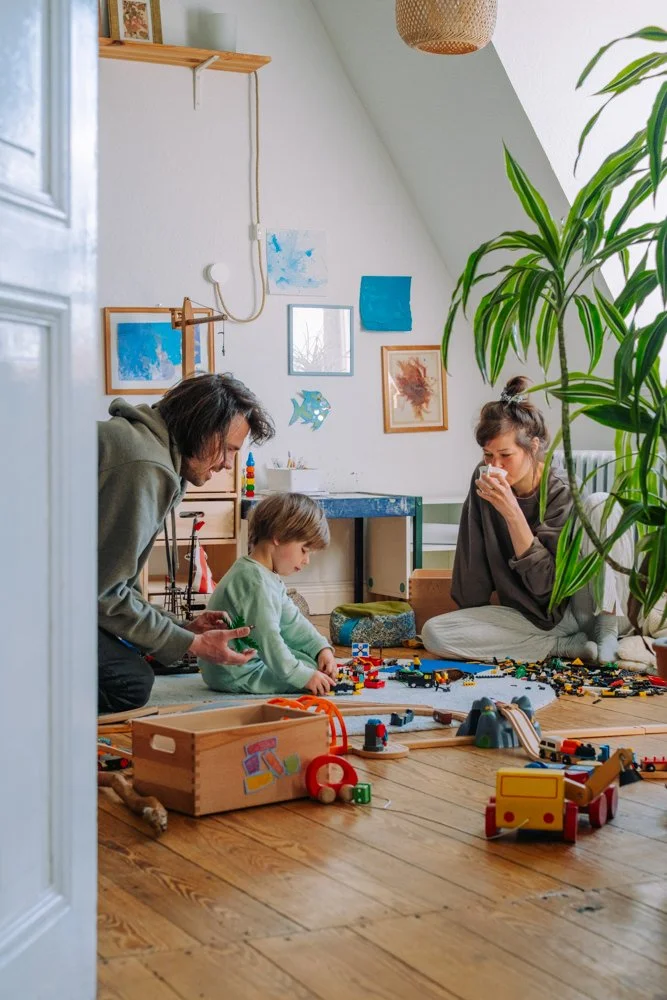 Familie spielt mit Spielzeug aus LEGO auf dem Holzfußboden in einem gemütlichen Zimmer mit Wandkunst und einem großen grünen Pflanze.