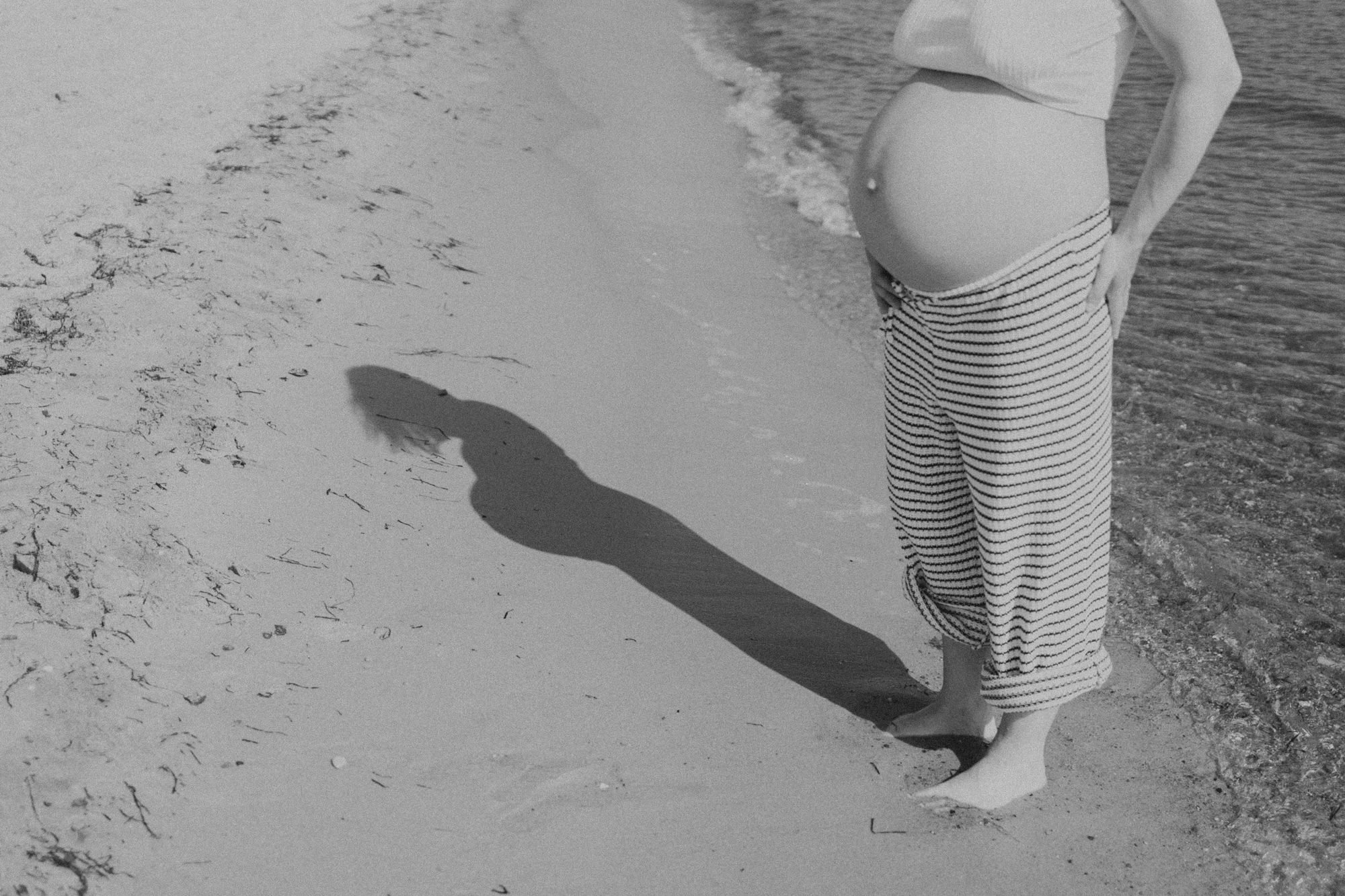 Schwangere Frau am Strand mit Schatten im Sand, bei Sonne, in lockerer Kleidung, barfuß
