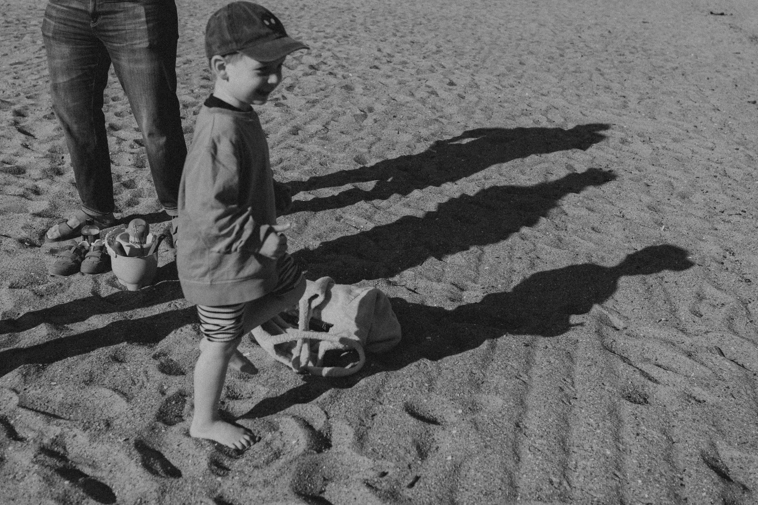 Ein Junge mit Mütze steht auf einem Sandstrand, neben ihm liegt ein Rucksack. Die Schatten der Person und des Rucksacks sind auf dem Sand sichtbar.