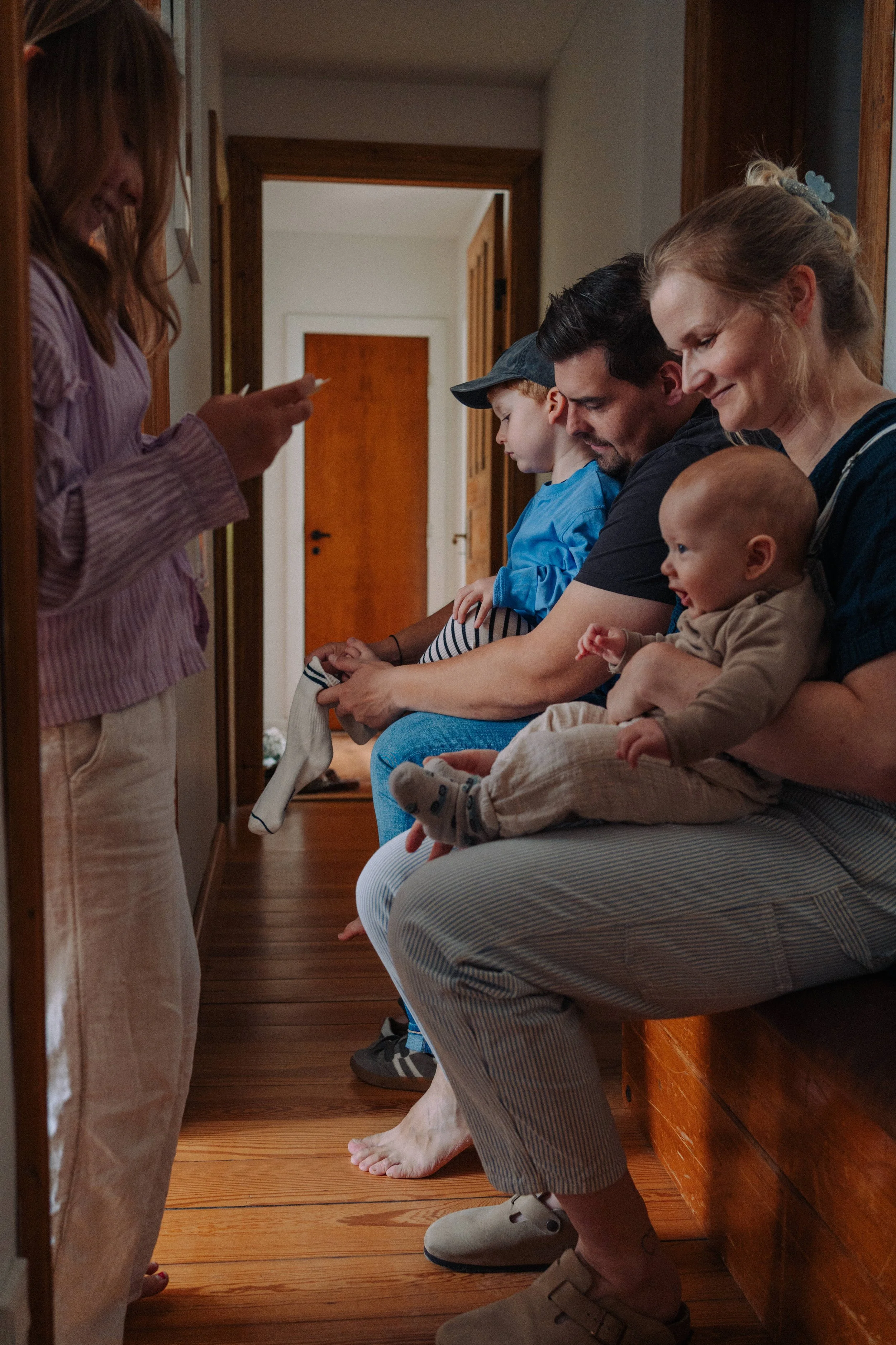 Familie sitzt auf einer Bank im Flur und spielt mit einem Baby, während ein Mädchen im Vordergrund ein Foto macht.