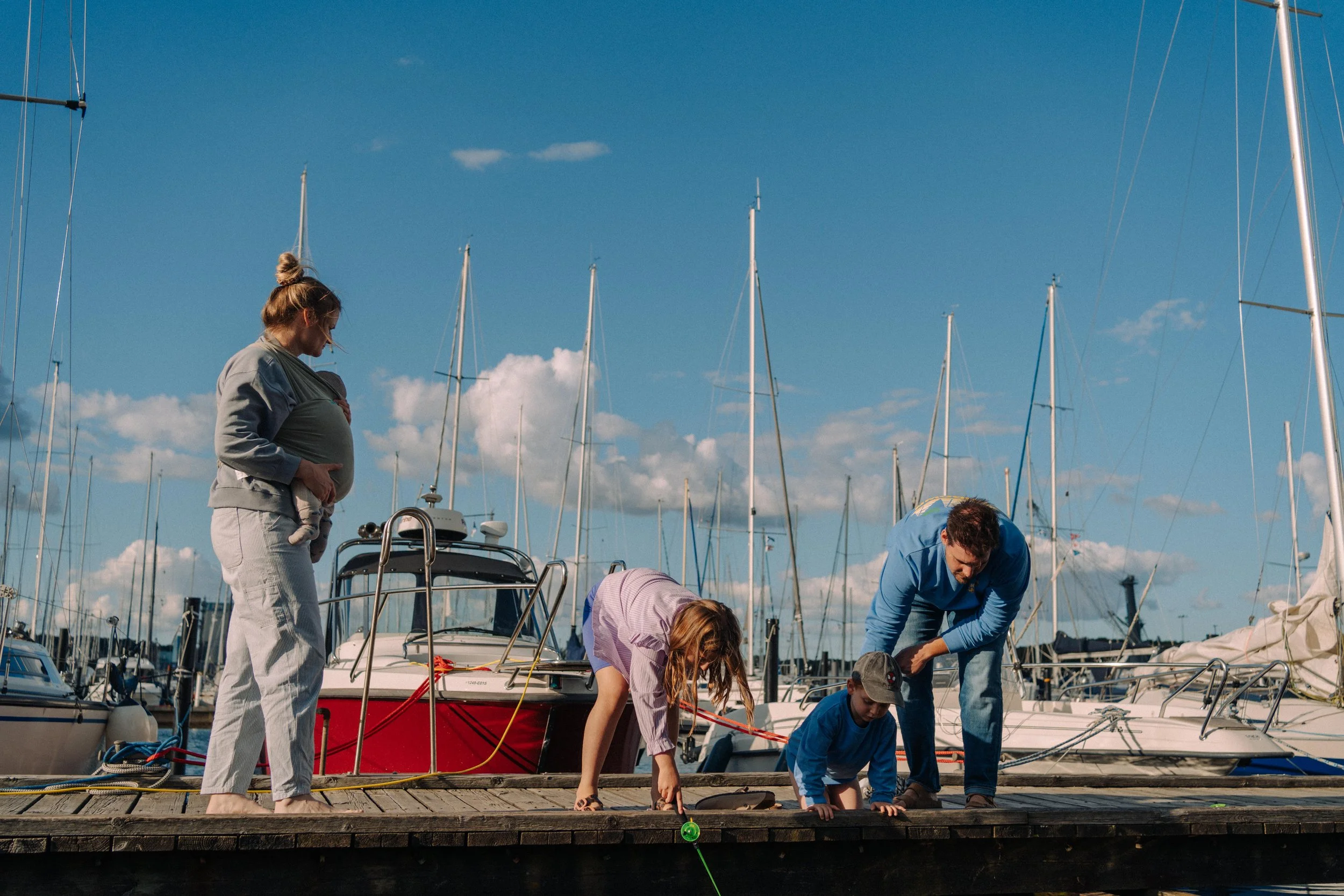 Familie auf einem Dock am Hafen beim Spaziergang mit Segelbooten im Hintergrund, sonniger Himmel.