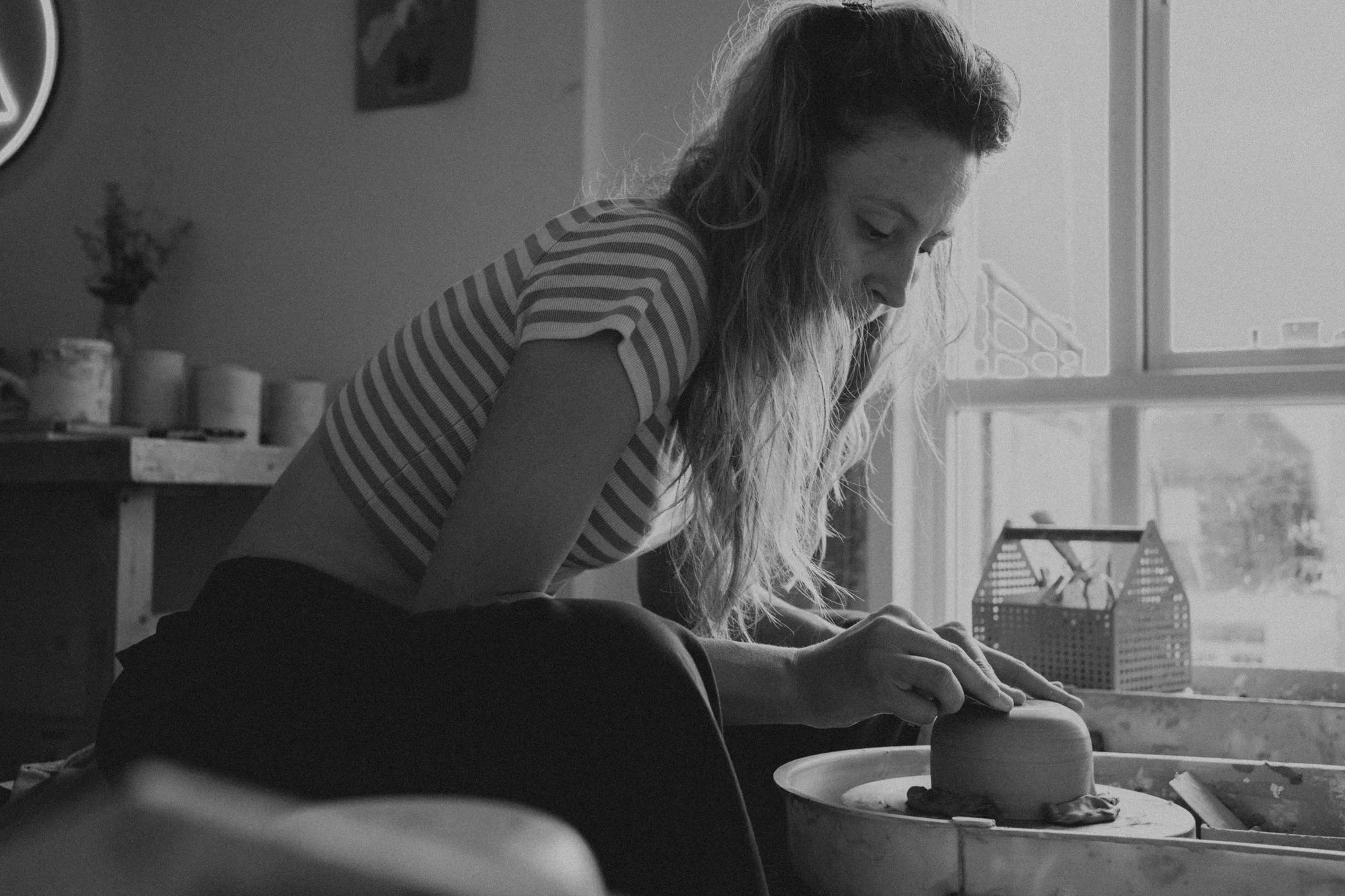 Eine junge Frau beim Töpfern in einem Atelier, arbeitet an einem Keramikstück auf einer Töpferscheibe neben einem Fenster.