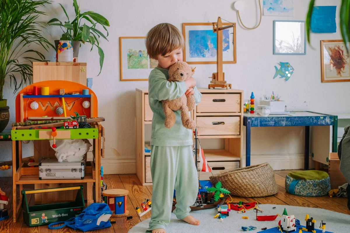 Ein Junge im Schlafanzug hält ein Stofftier in einem Kinderzimmer. Das Zimmer ist mit Spielsachen, bunten Bildern und Dekorationen gefüllt.