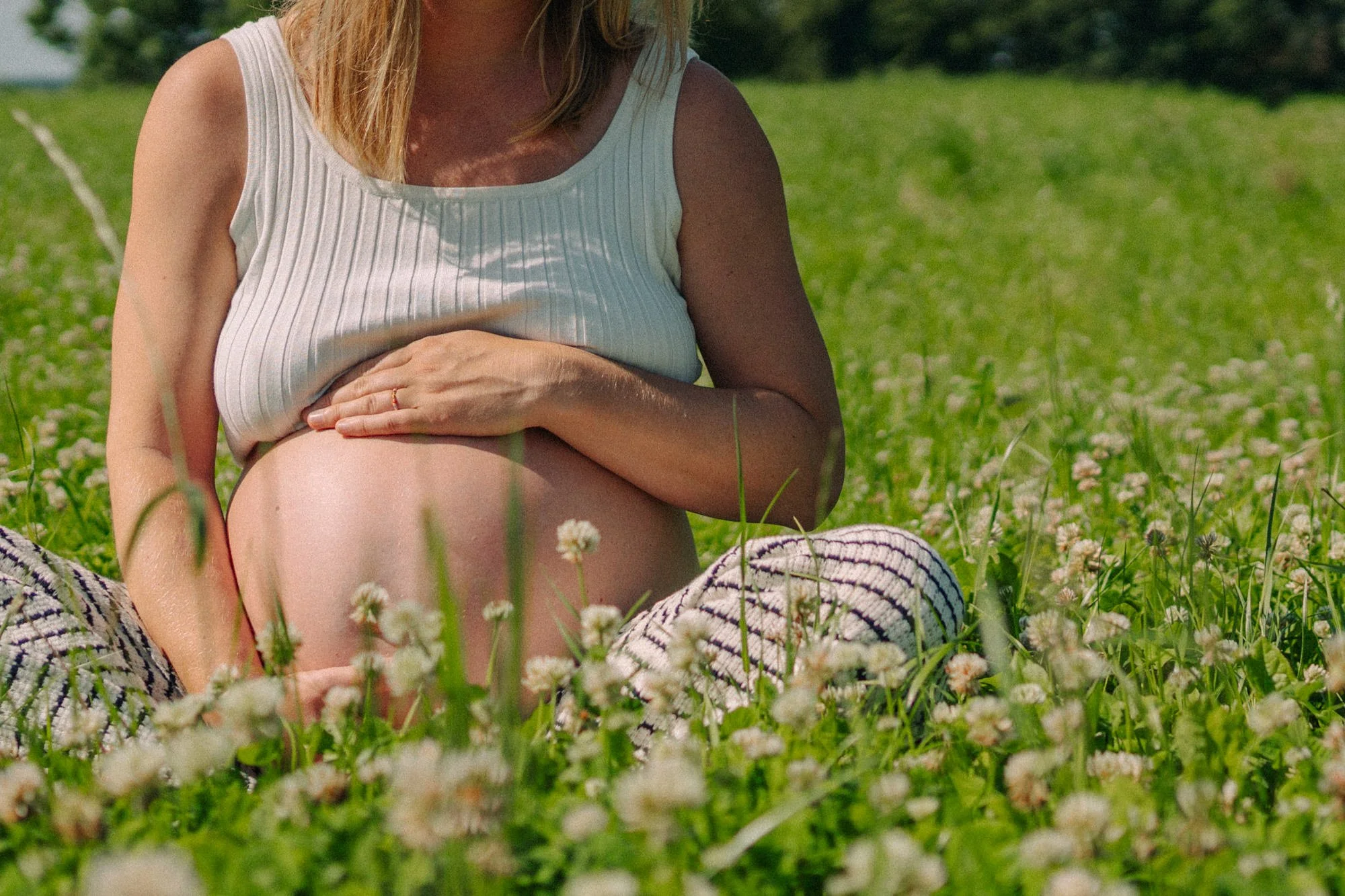 Schwangere Frau sitzt im Grünen auf einer Wiese mit weißen Blumen, trägt ein hellgraues ärmelloses Top und hat die Hand auf den Bauch gelegt.