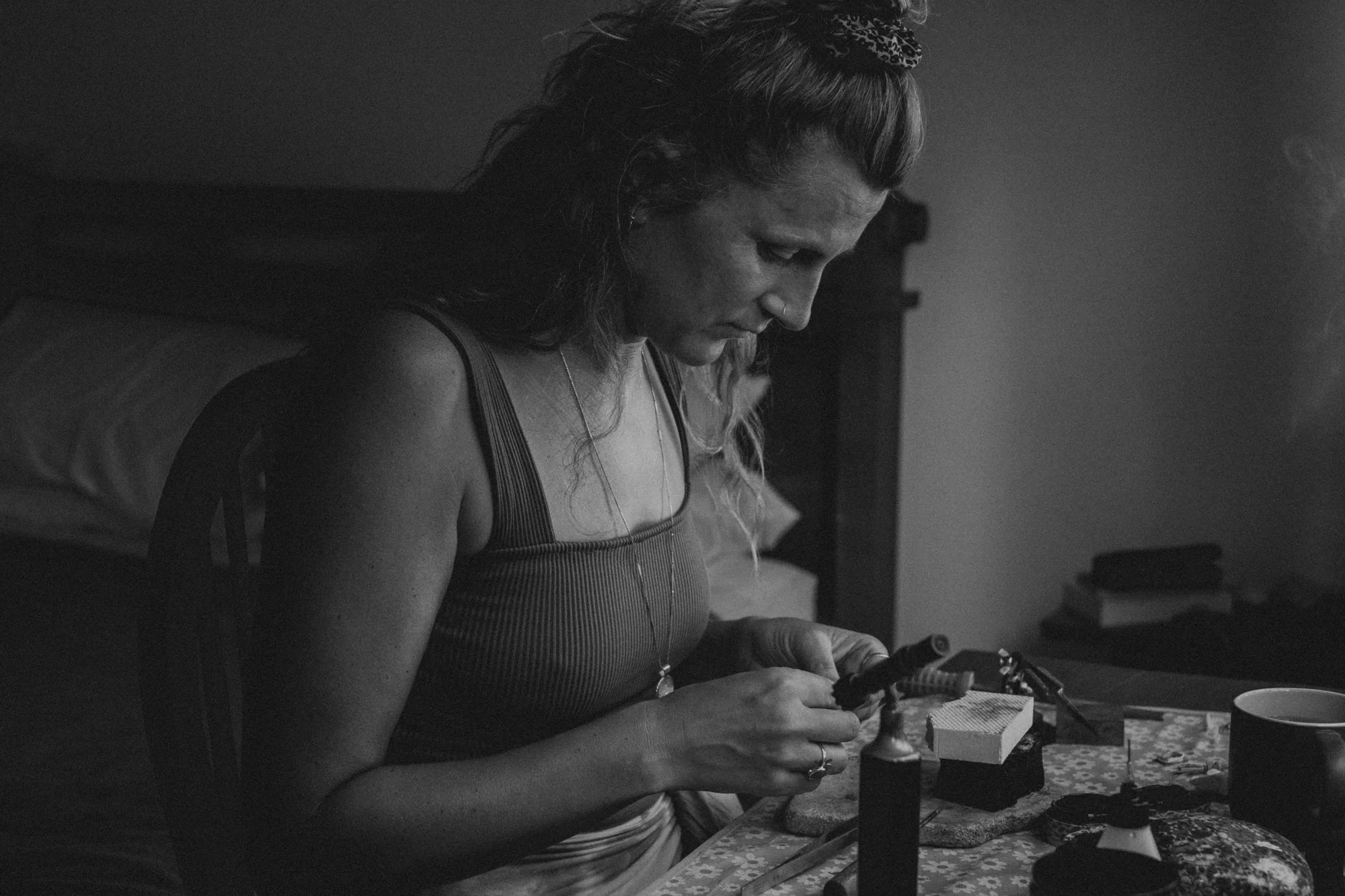 Schwarz-Weiß-Foto einer Frau beim Tätowieren zu Hause. Sie sitzt an einem Tisch mit Tätowierwerkzeug, definierte Konzentration auf die Arbeit. Im Hintergrund ein Bett und Bücher auf einem Nachttisch.