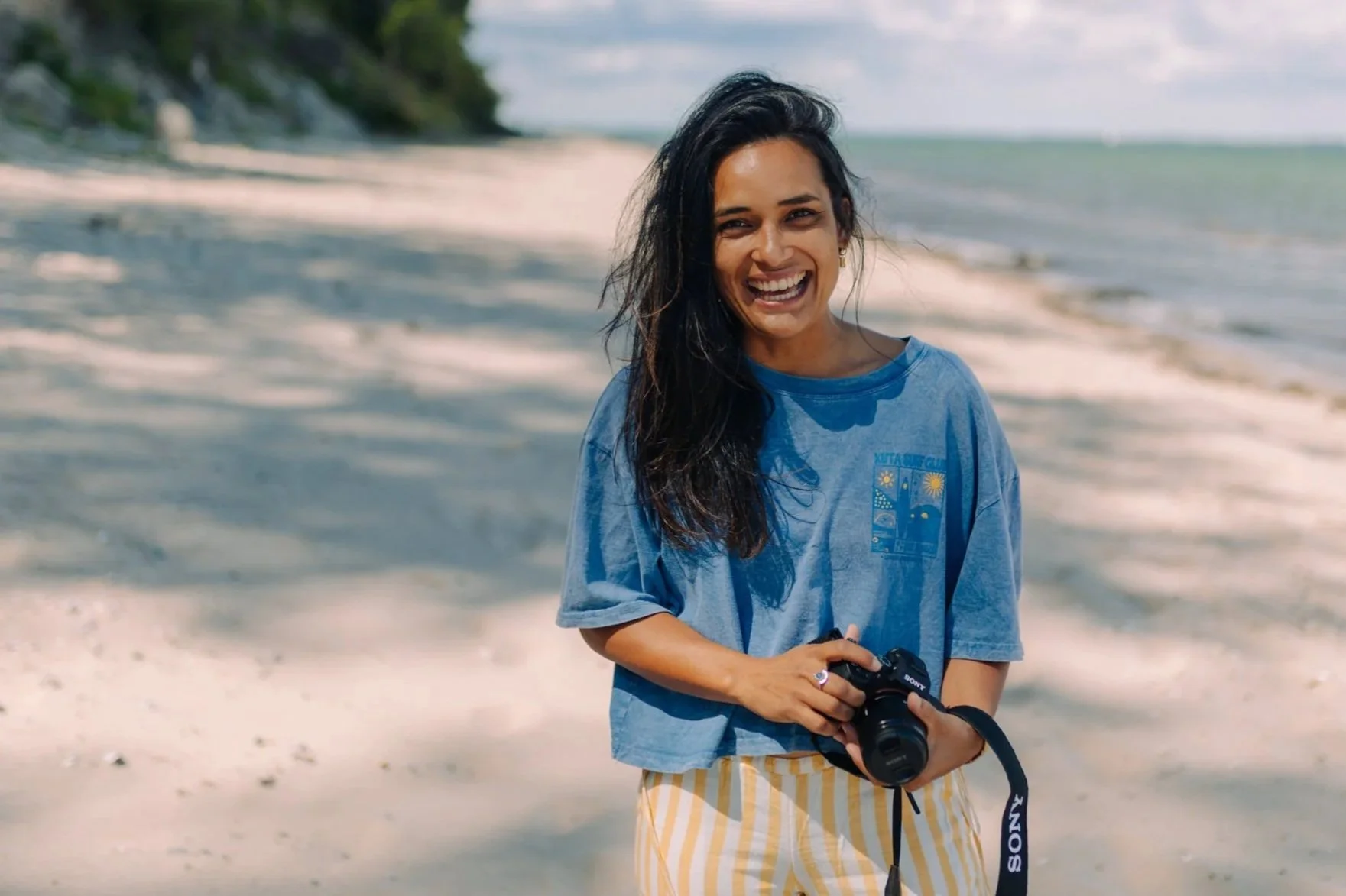 Eine lächelnde Frau mit schwarzen, lockigen Haaren steht am Strand und hält eine Kamera in der Hand.