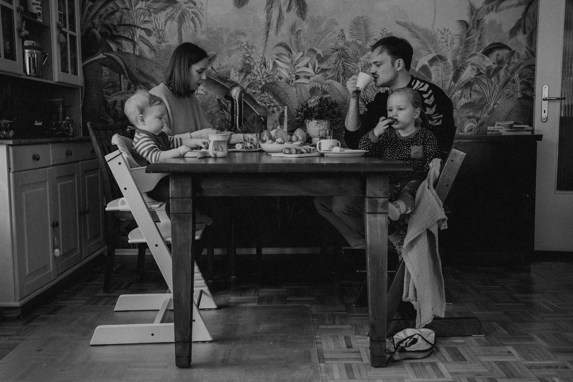 Familie sitzt beim Frühstück an einem Holztisch in einer Küche mit pflanzenbezogener Tapete im Hintergrund, Kinder essen, Erwachsene trinken Kaffee.