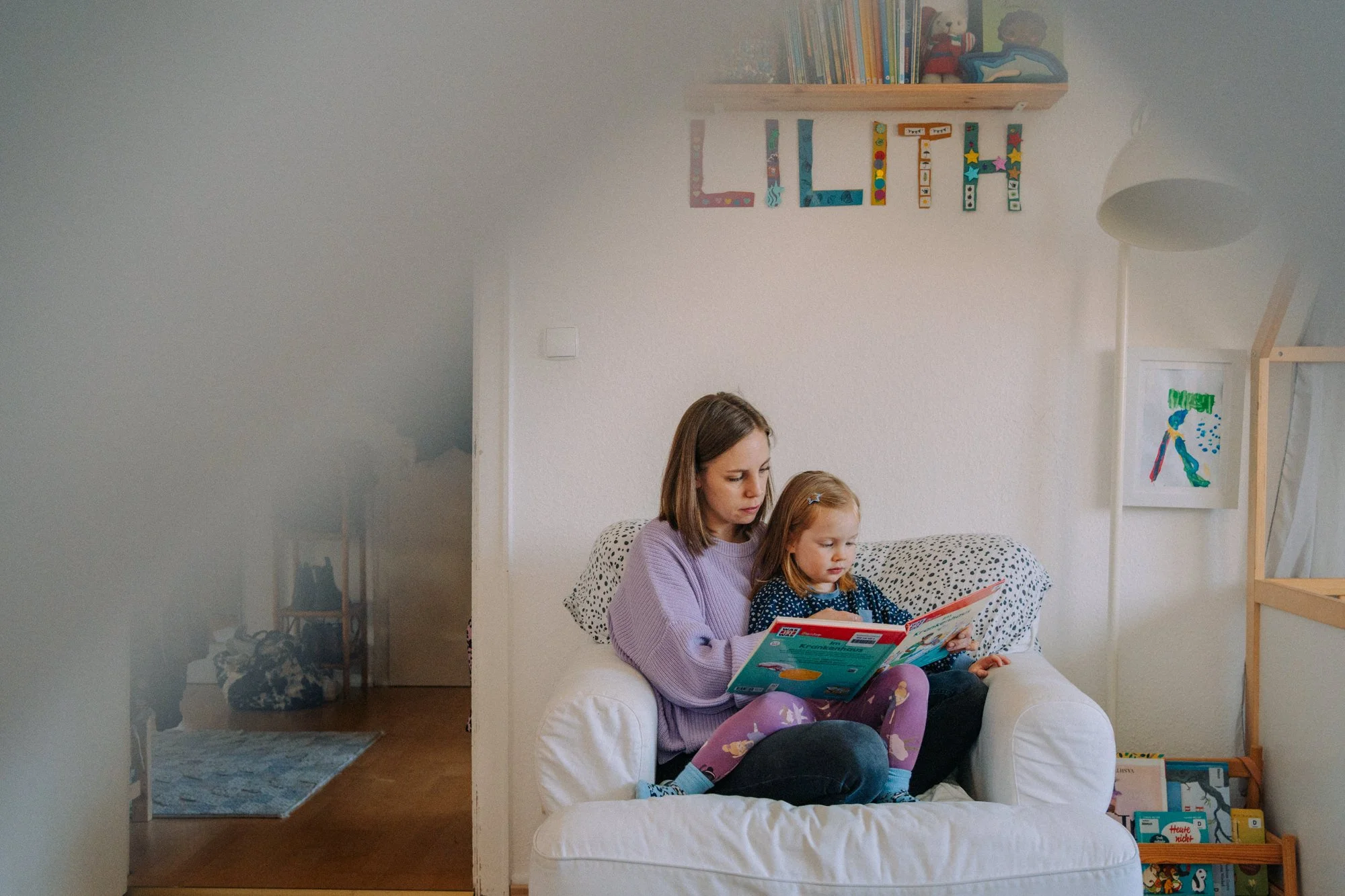 Eine Frau und ein Mädchen sitzen zusammen auf einem weißen Sessel, lesen ein Buch und scheinen konzentriert zu sein, in einem hellen, gemütlichen Wohnzimmer.